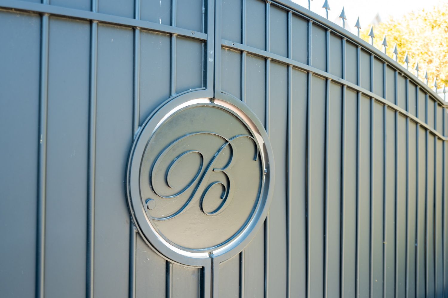 Elegant gray metal gate featuring a decorative circular medallion with ornate letter B monogram in script font.