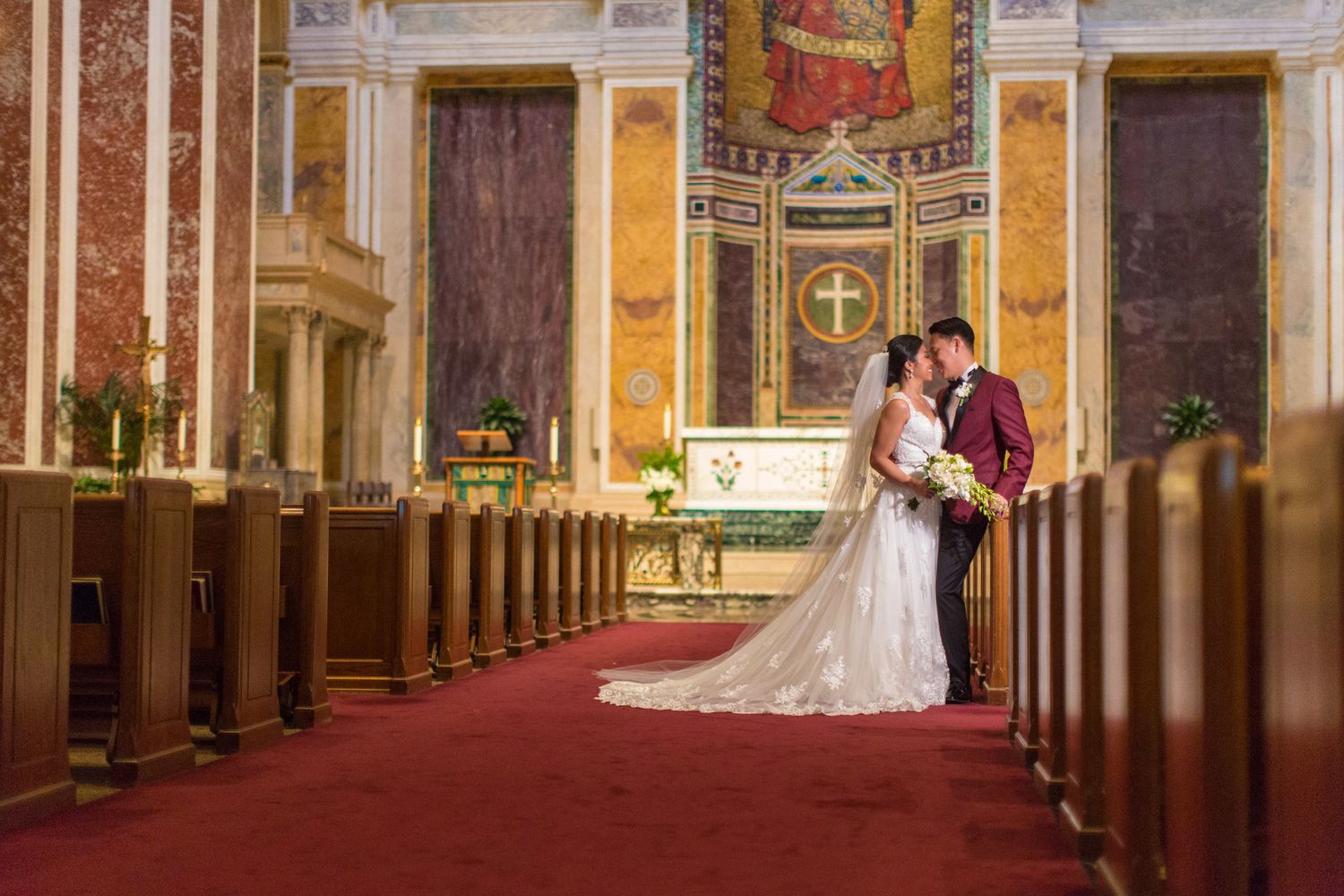Bride and groom share a romantic kiss at the altar of an ornate church with red carpet aisle.