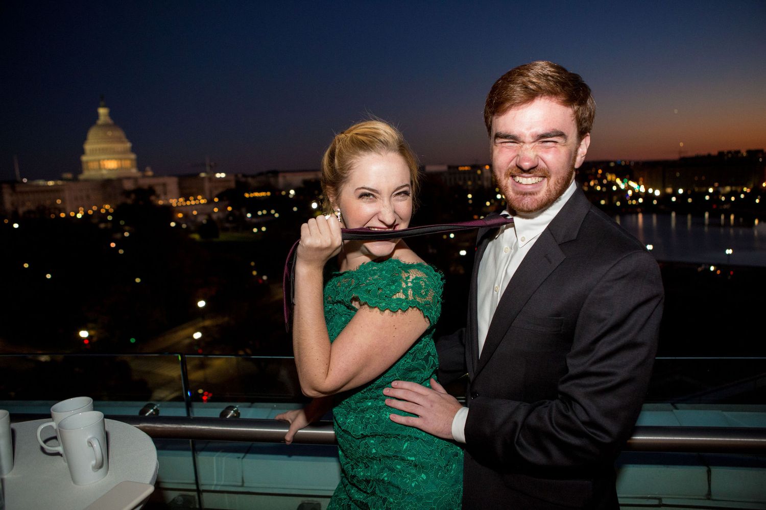 Couple posing on rooftop terrace at dusk with Capitol building backdrop, woman in green lace dress holding fake mustache.