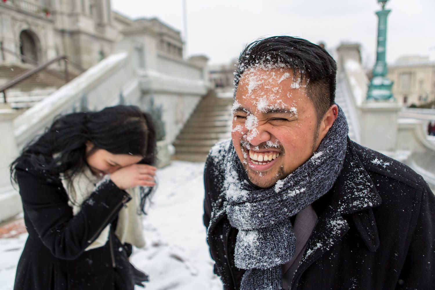 Two people enjoying a snowball fight outdoors in winter, man laughing with snow on his face and scarf.