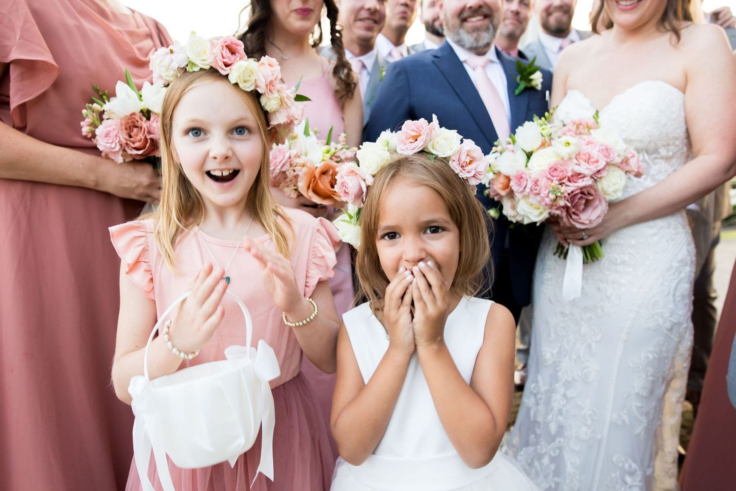 Two flower girls in pink and white dresses with floral crowns react with joy and surprise at a wedding ceremony.