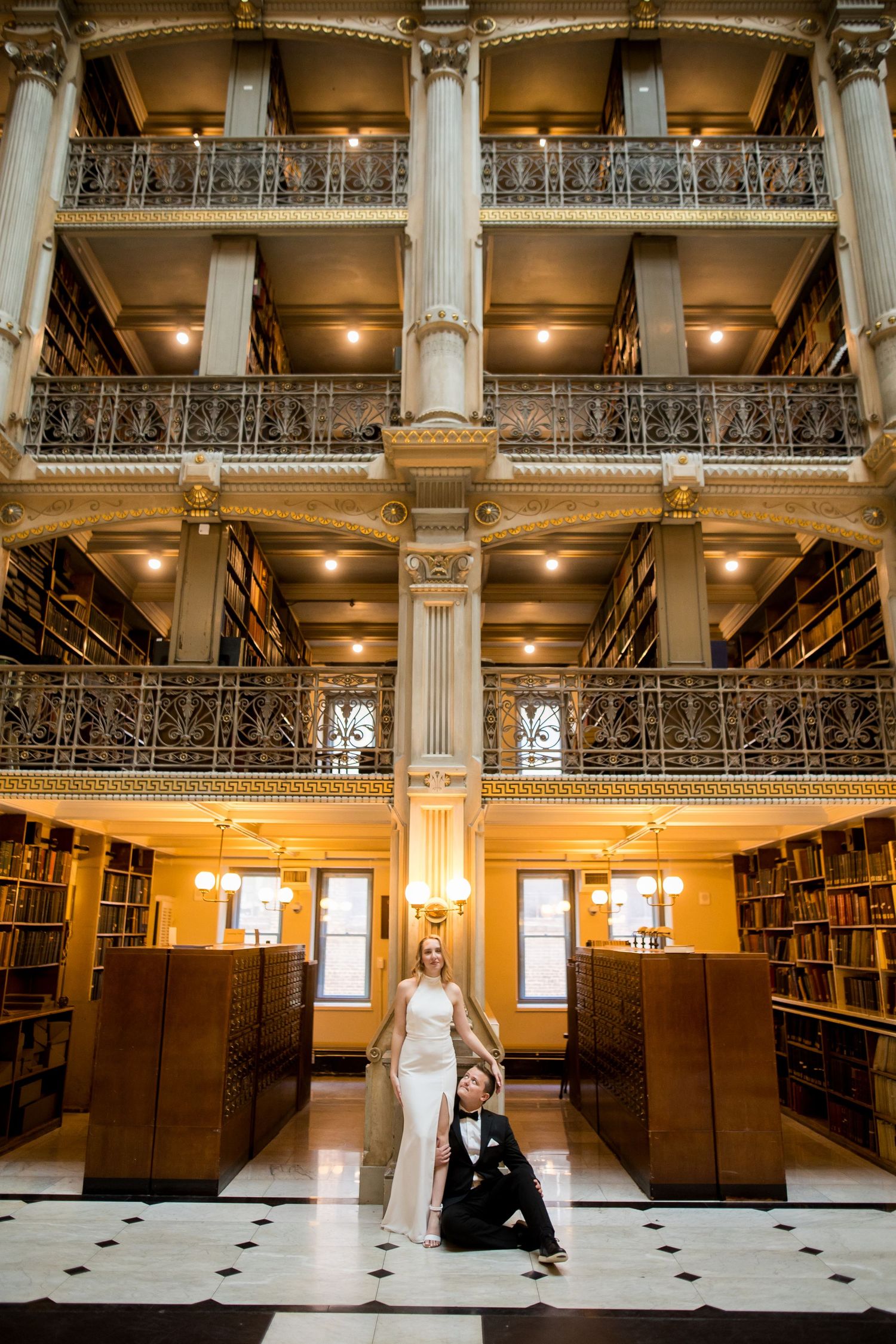 Bride and groom posing in the George Peabody Library with ornate iron balconies and warm lighting in Baltimore, MD.