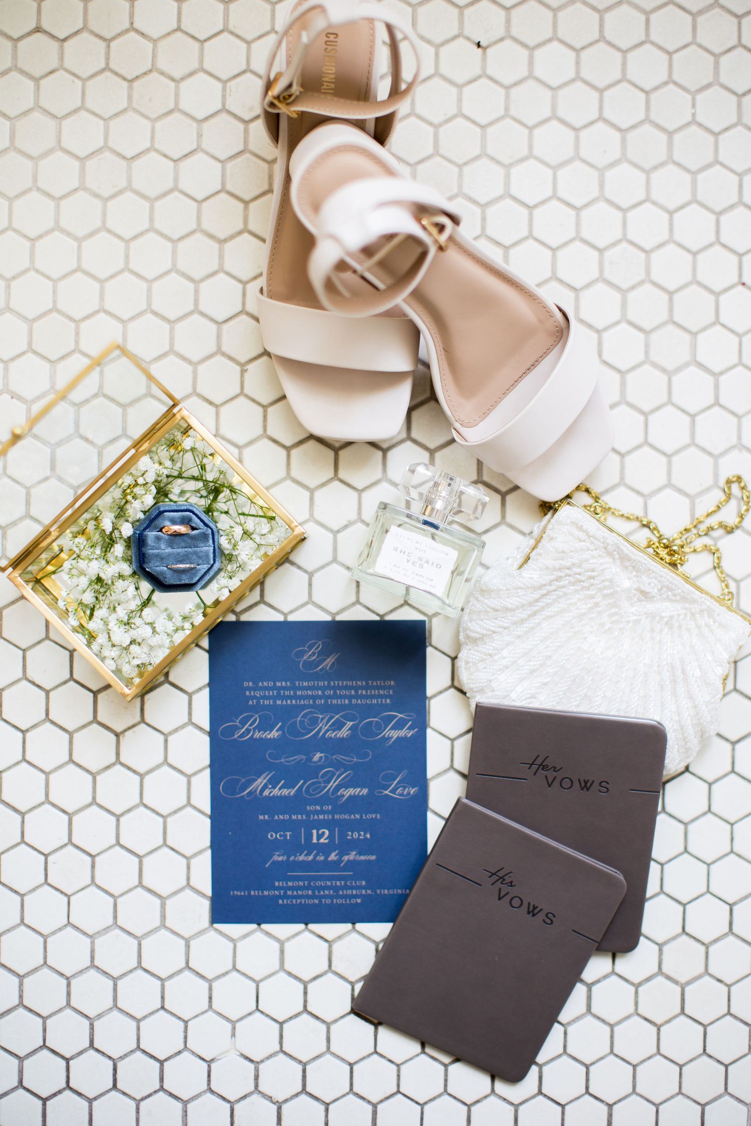 Elegant wedding flat lay with navy invitation, nude heels, vow books, perfume, and floral ring box on hexagon tile.
