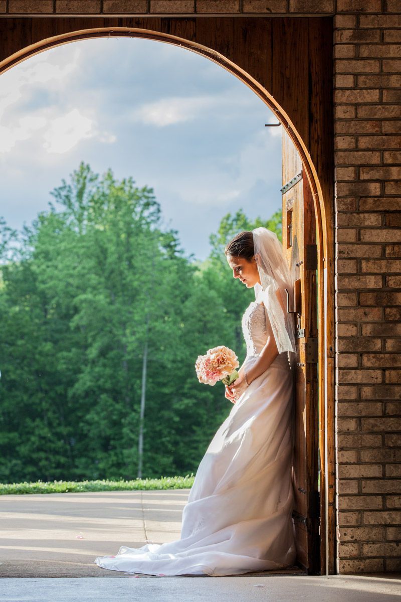 Bride in white gown and veil holds pink bouquet in arched wooden church doorway with green trees backdrop.