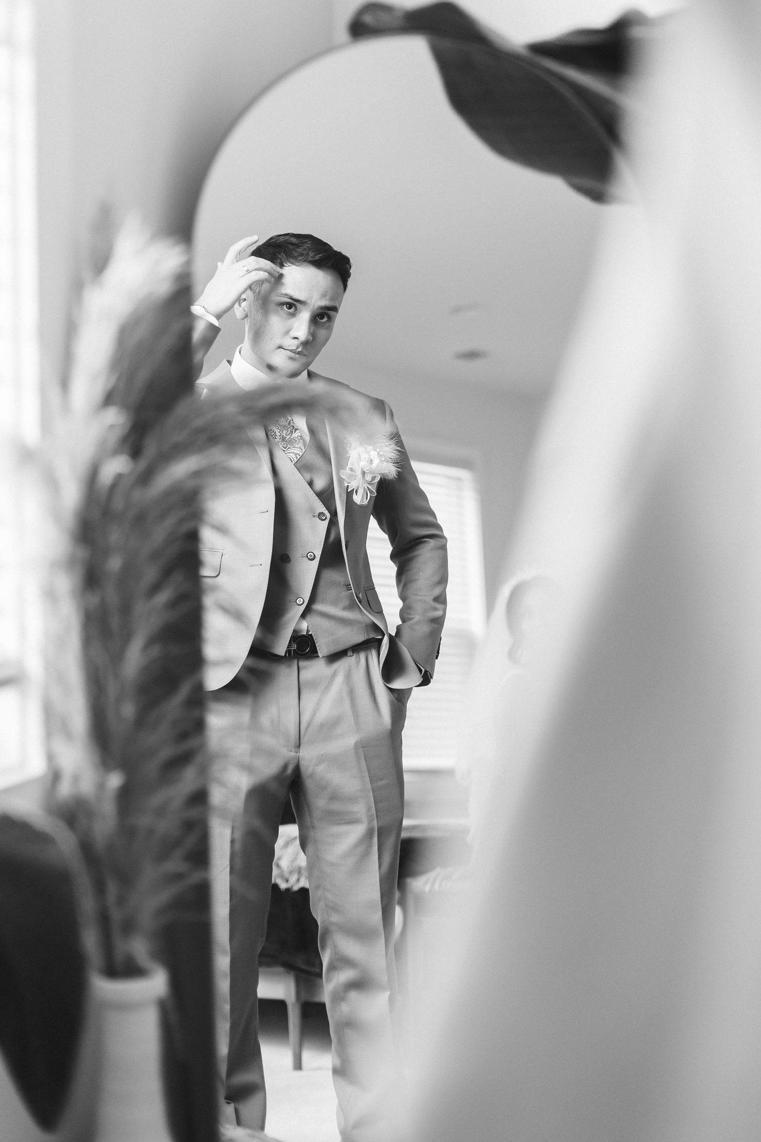 Black and white photo of a groom in a suit adjusting his hair while checking his reflection in a mirror.