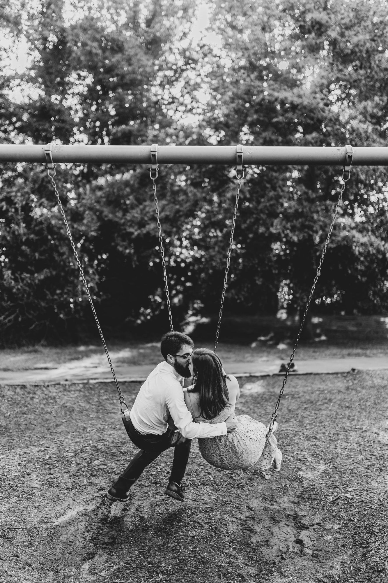 Black and white photo of a couple sharing a kiss on a playground swing set surrounded by trees.