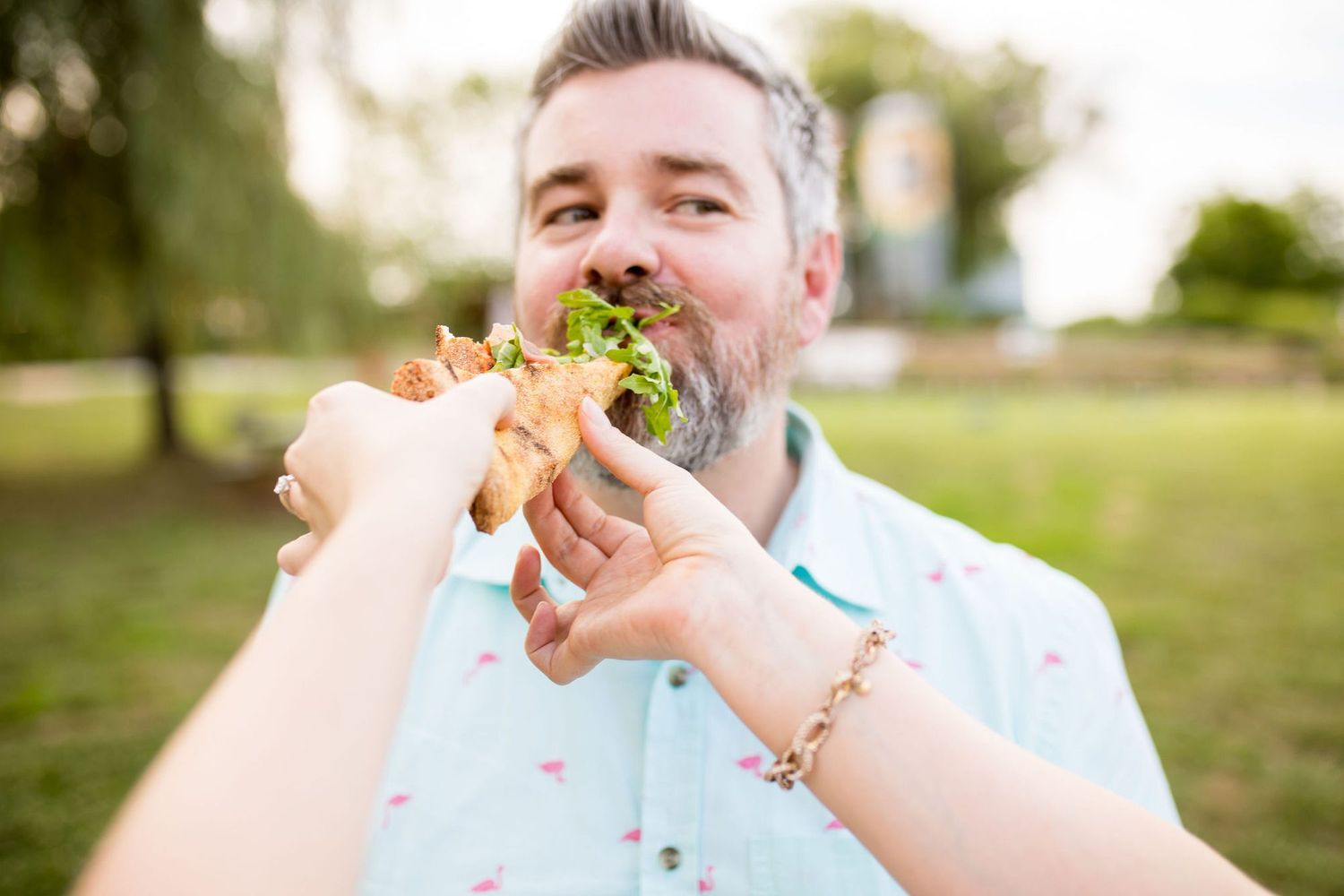 Man with gray beard eating a large sandwich outdoors in a park on a sunny day.