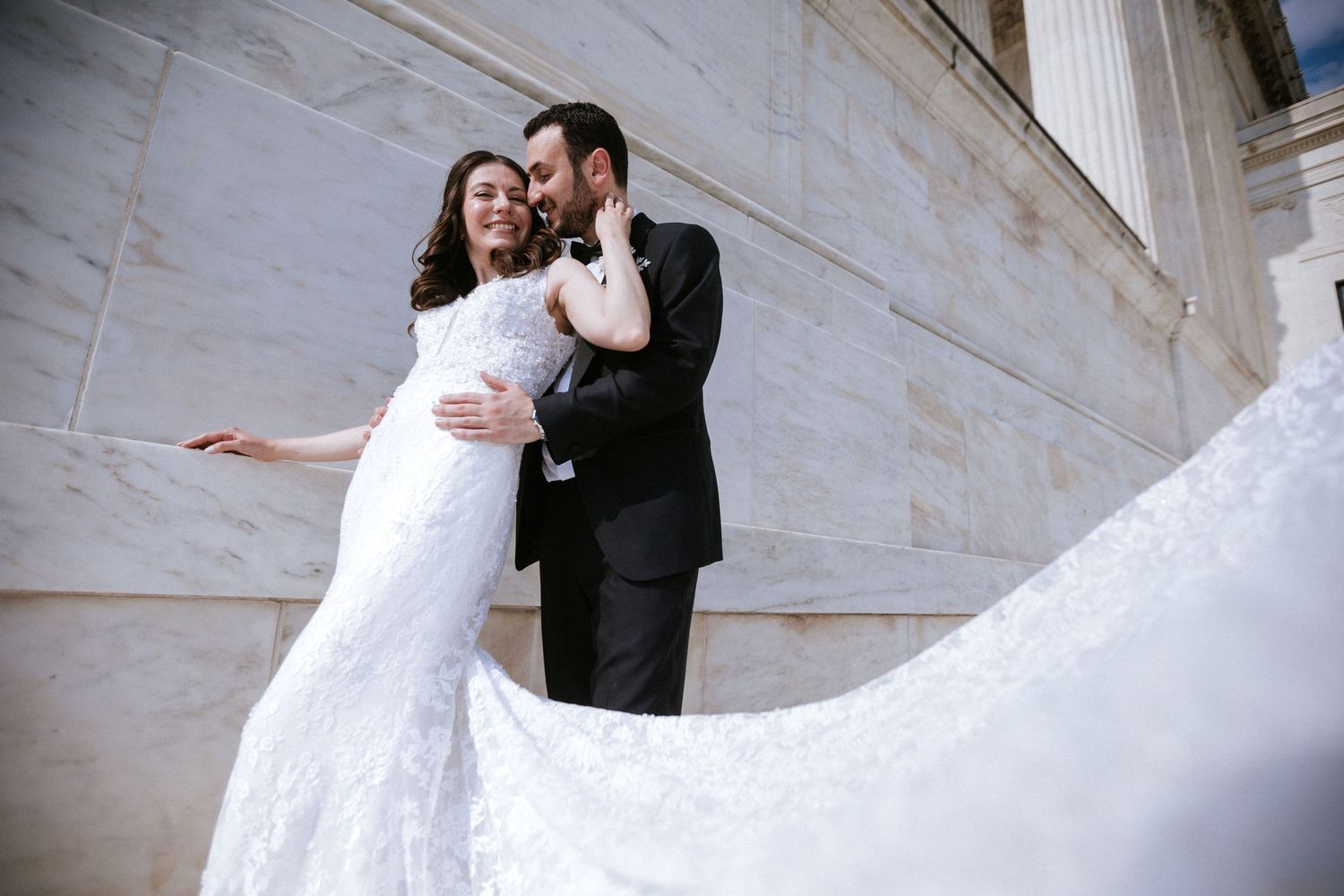 Bride in white lace gown and groom in black tuxedo embrace on marble steps in elegant wedding photo.