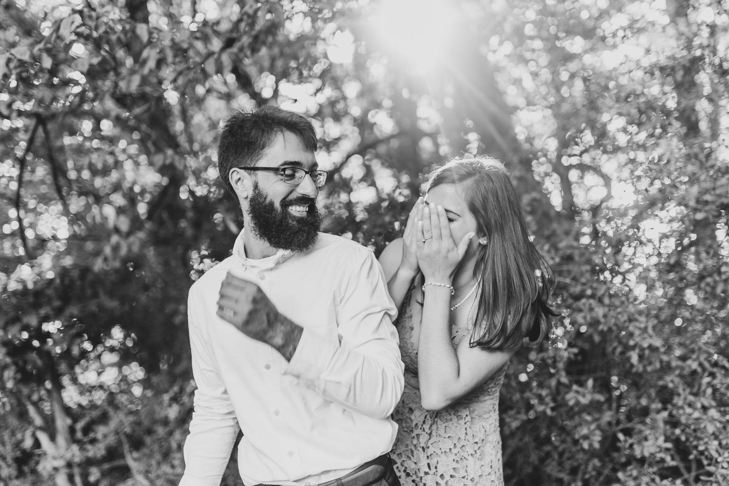 Black and white photo of a bearded man laughing with a woman covering her face with her hands outdoors.