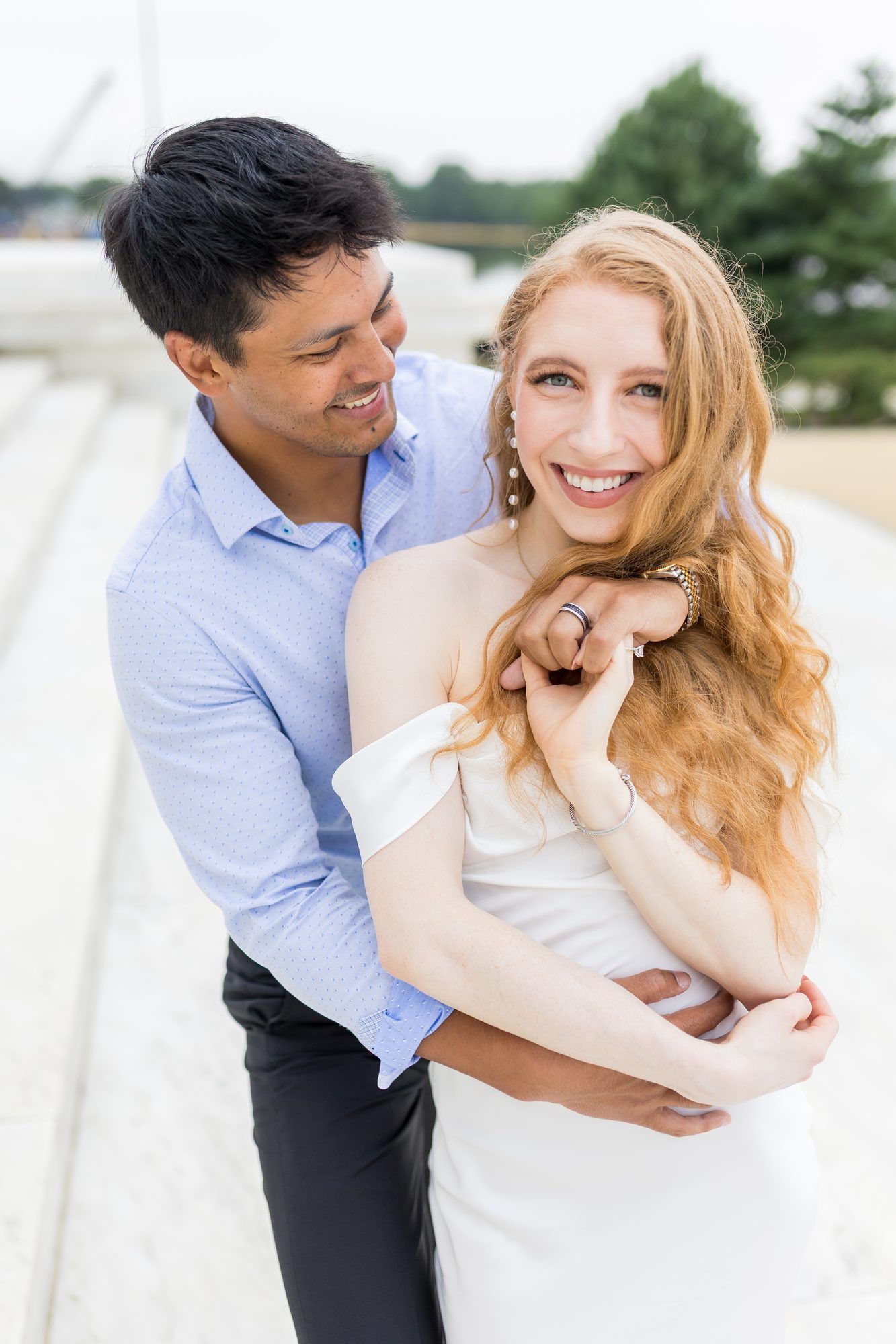 Happy couple embracing outdoors, smiling woman in white dress with red hair, man in blue shirt hugging her from behind.
