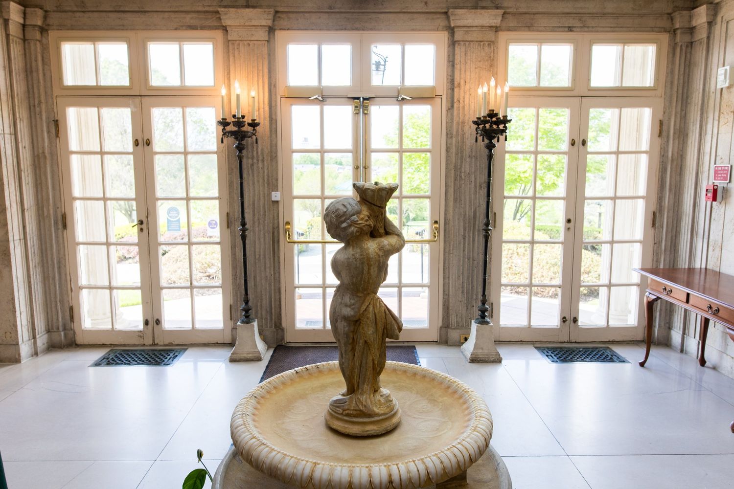 Elegant indoor fountain with classical female statue in a grand hall featuring tall French doors and ornate columns.