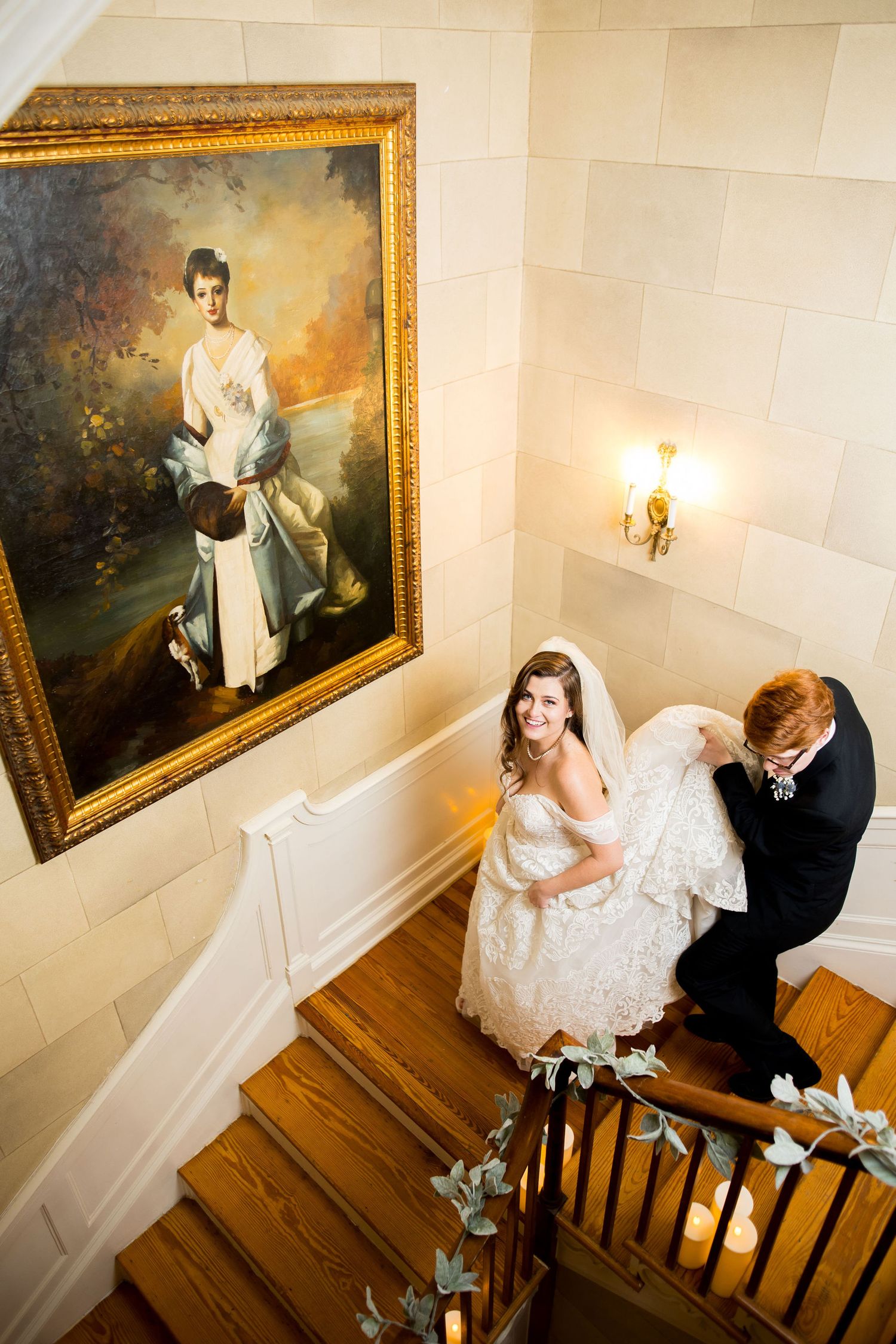 Bride and groom on elegant staircase with classic portrait, groom lifting wedding dress train in grand venue.