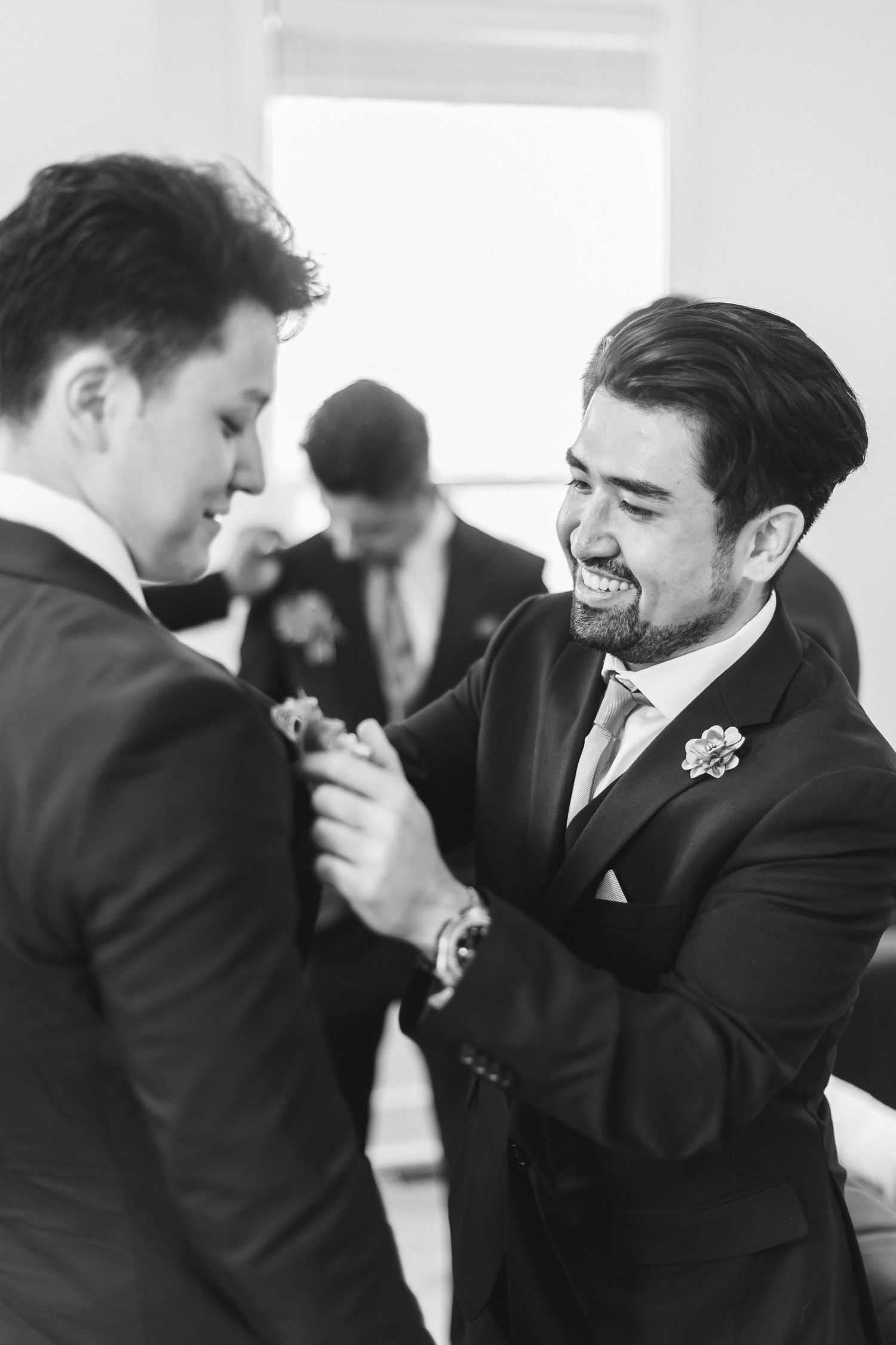 Black and white photo of two grooms in suits smiling while pinning boutonnieres at a wedding.