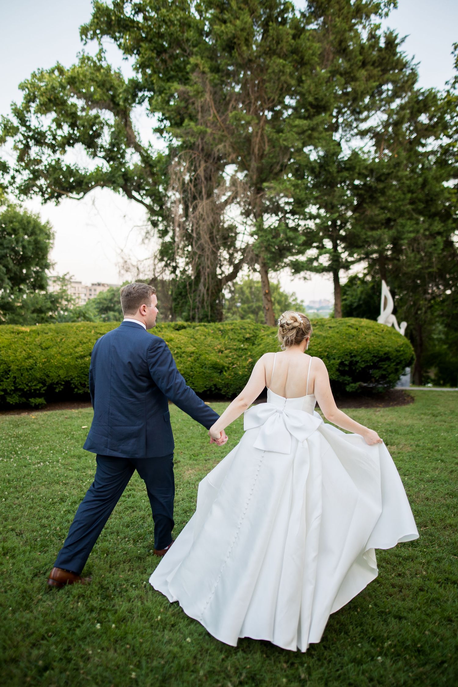 Bride and groom walking hand in hand through lush green garden, bride in elegant white ballgown, groom in navy suit.