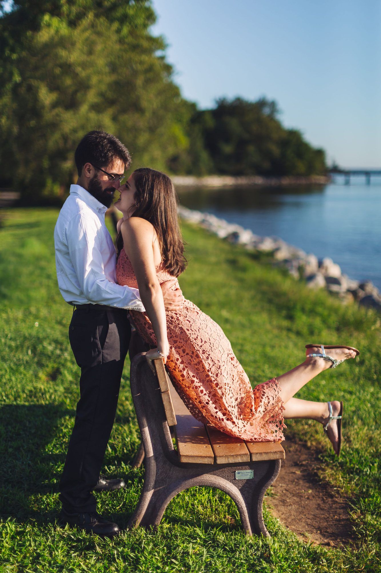 Couple sharing a romantic kiss on a park bench by the waterfront, woman in pink lace dress, sunny day.