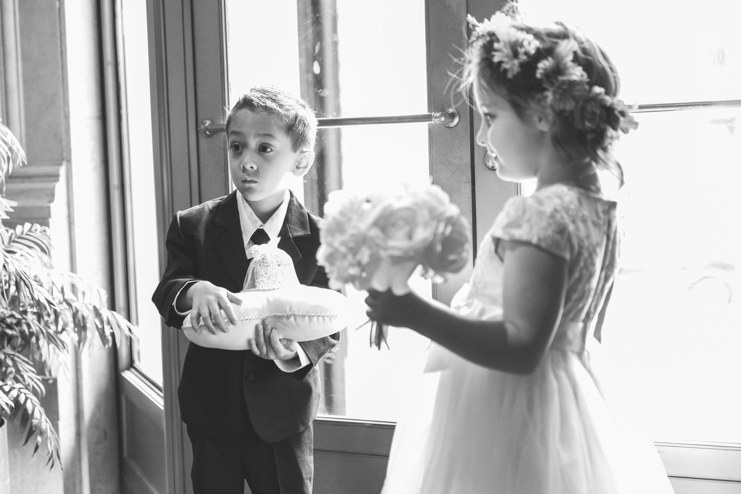 Black and white photo of a young ring bearer and flower girl standing by a window during a wedding ceremony.