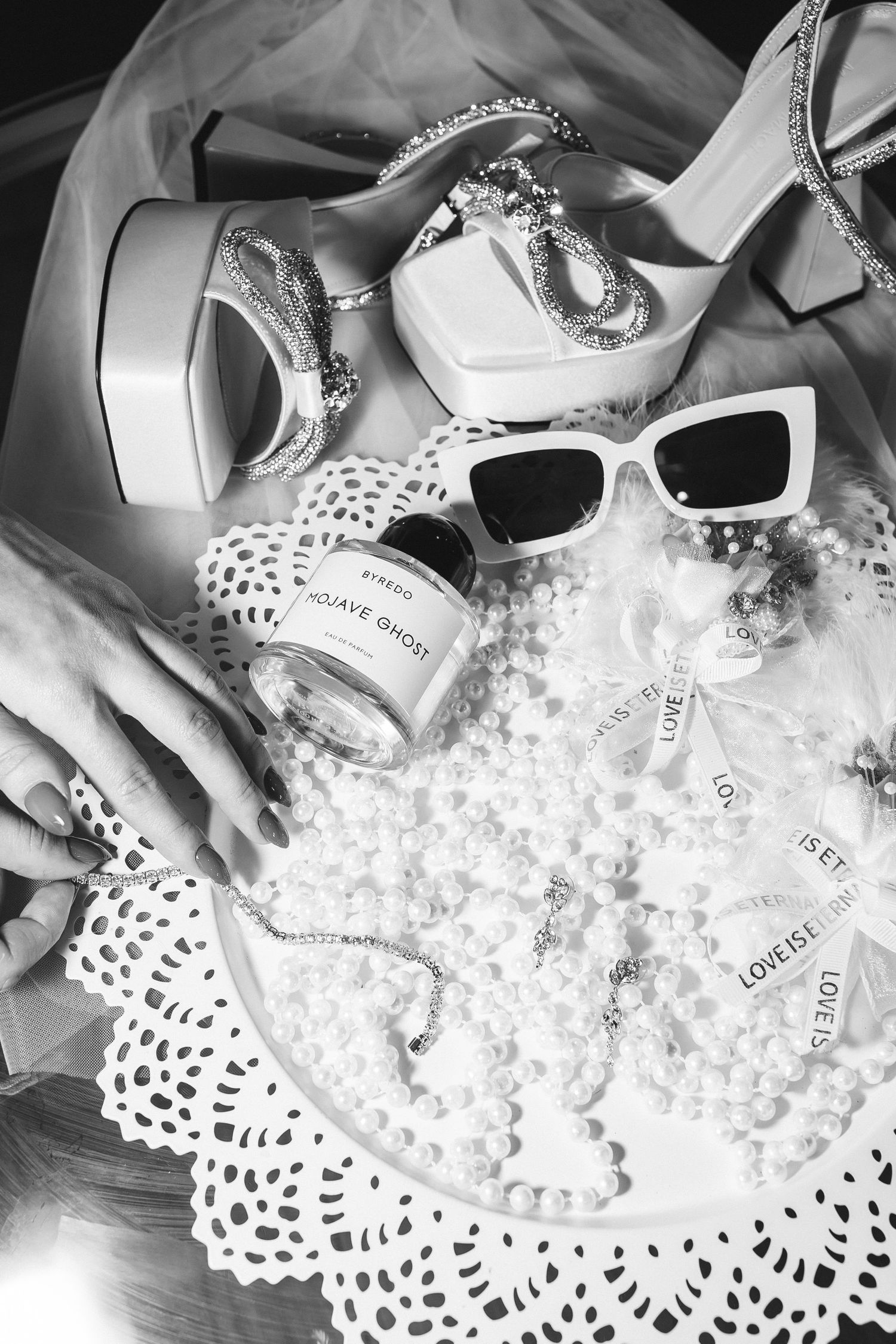 Black and white flat lay of bridal accessories including heels, pearls, sunglasses, and perfume on lace doily.
