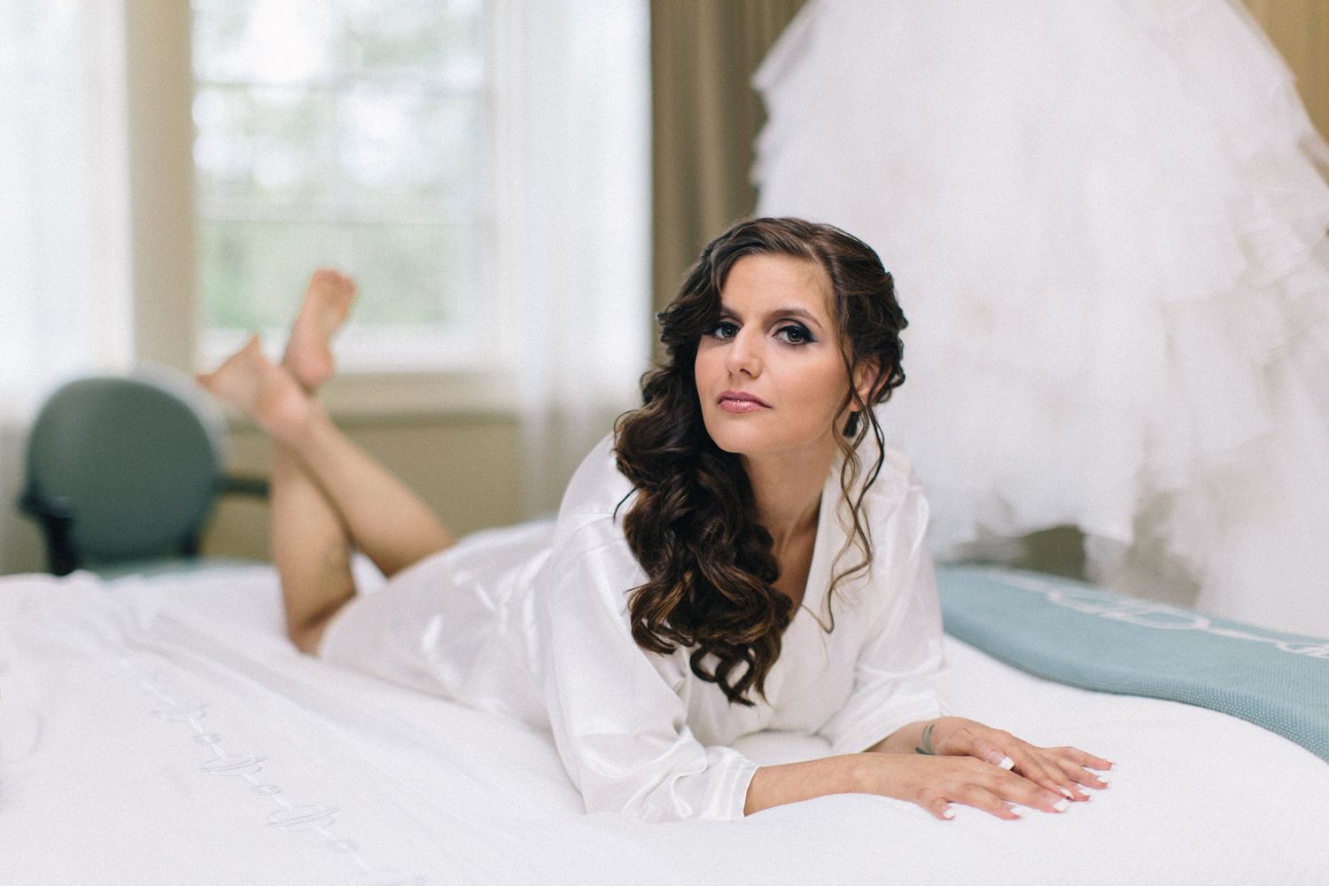 Elegant bride in white satin robe lying on bed with wedding dress in background during bridal boudoir session.