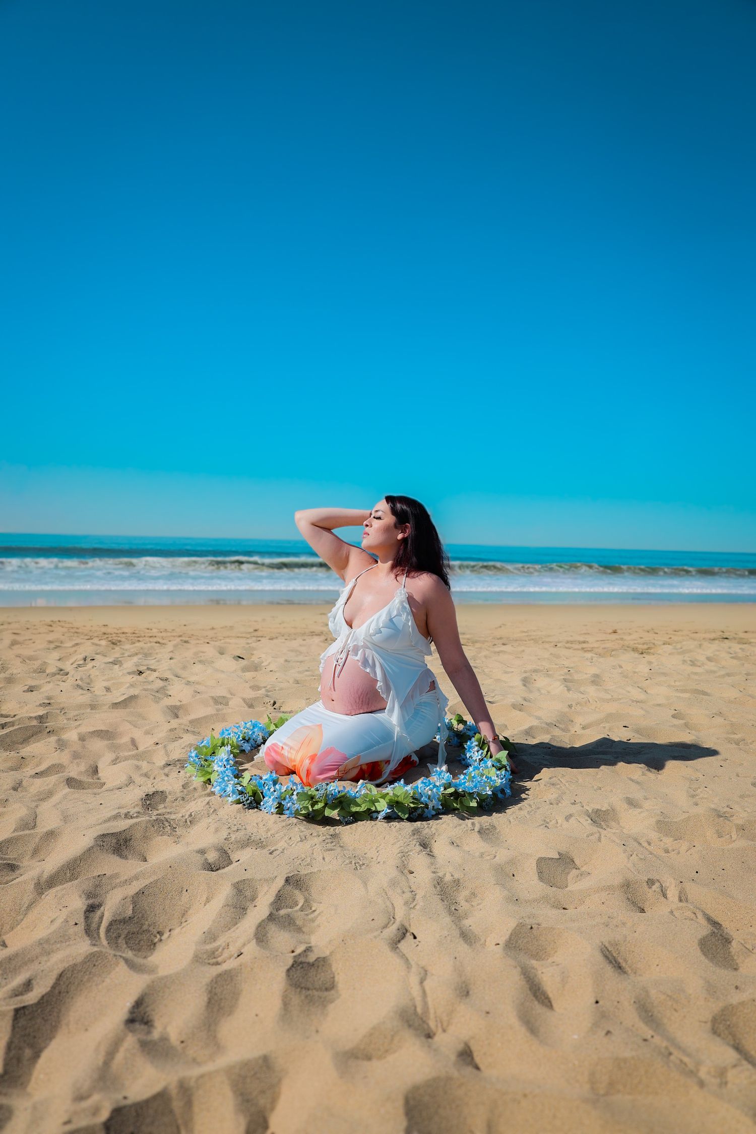 Kneeling maternity portrait of expecting mom in white outfit, surrounded by a delicate blue floral garland on a sunny beach.