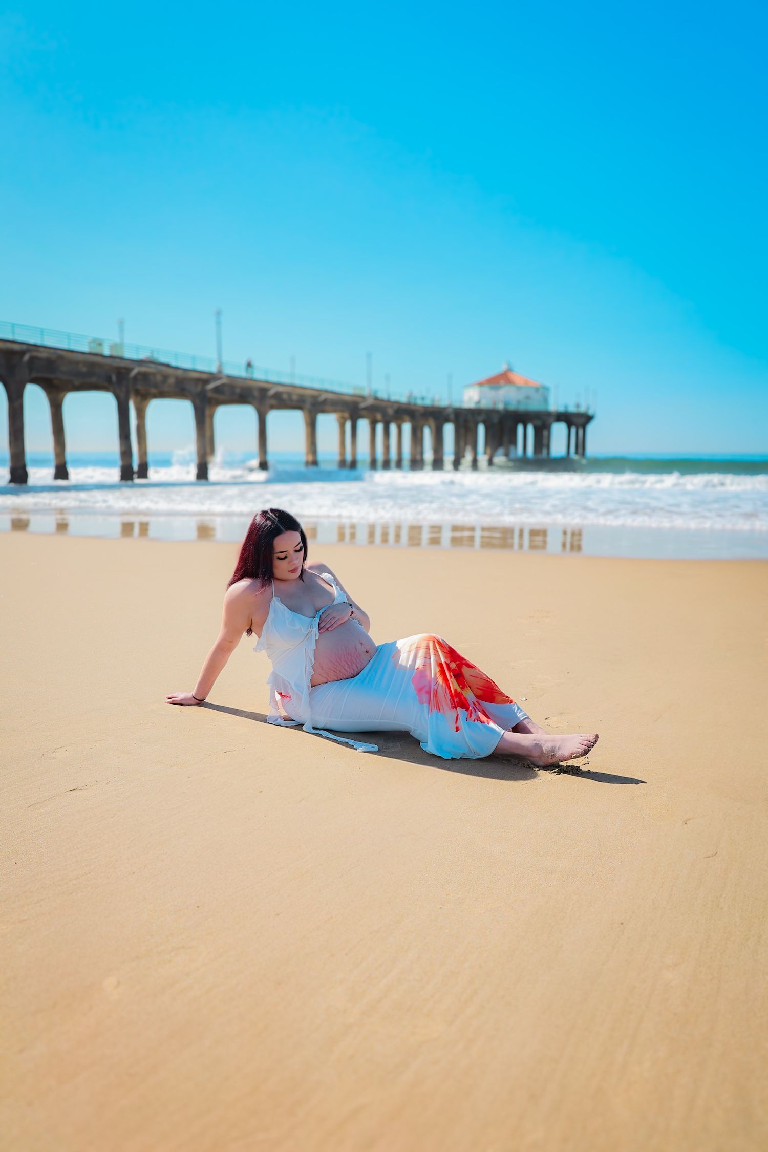 Pregnant mom seated on the sand in white and orange floral attire, gently cradling her belly with a serene expression.