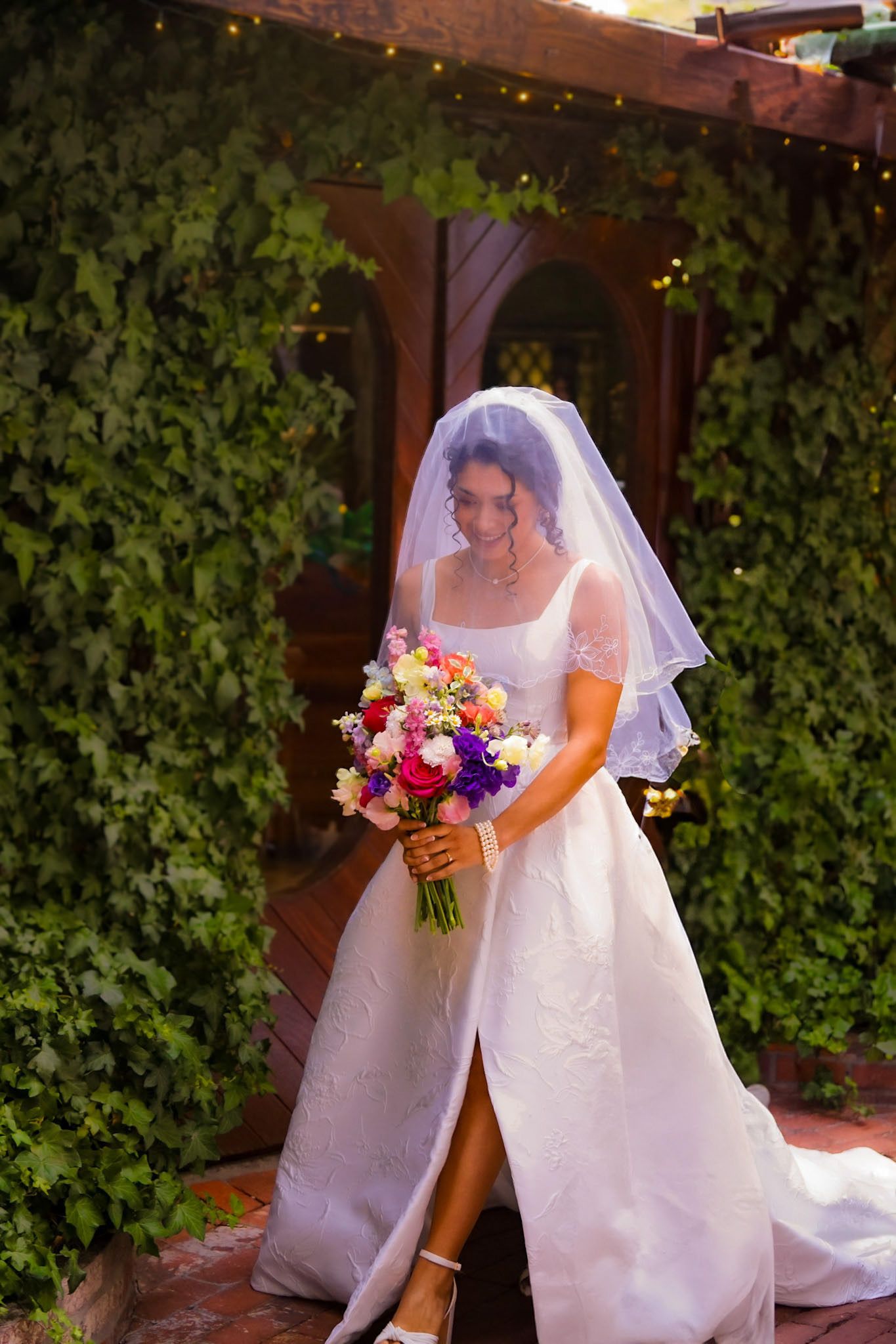 The bride walks towards the entrance to meet her parents to walk her down the aisle, veil over her head, bouquet in hand and her dress flows