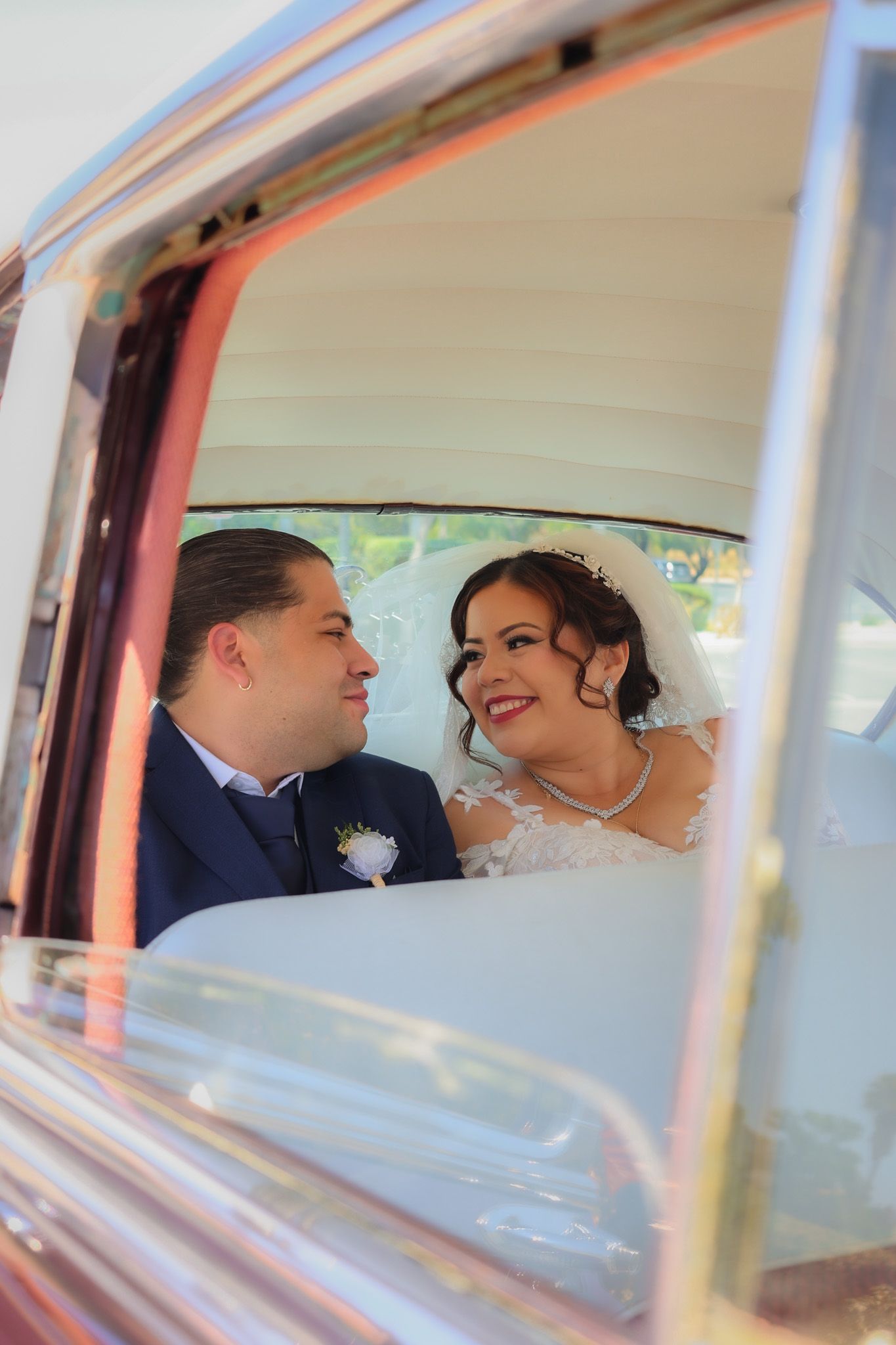 Newlywed couple smiling, holding hands while seated in a vintage classic car after their wedding ceremony, sunlight reflecting off the car.