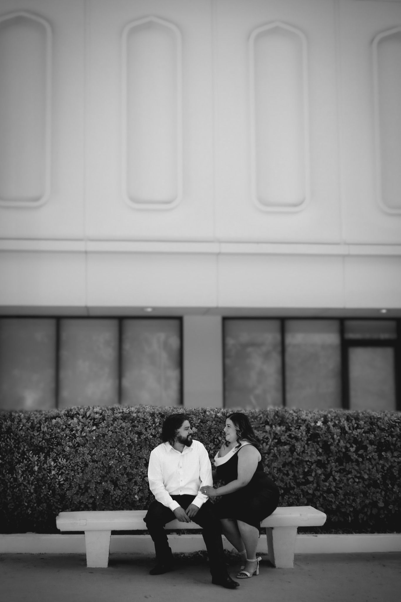 Engagement session for couple sitting on a bench outdoors, facing one another with a beautiful white building and hedges behind them.