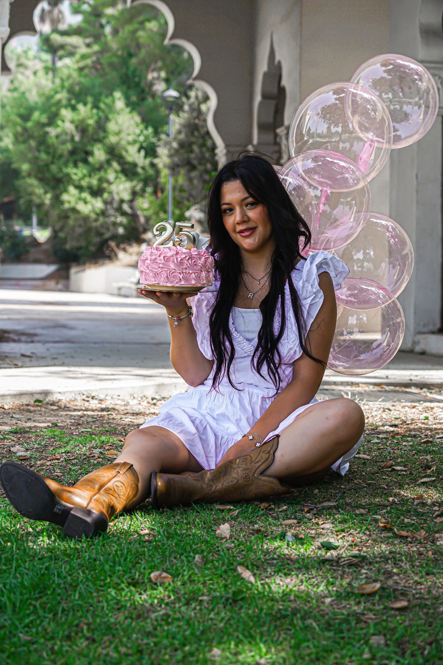 Woman sitting in front of a white library with beautiful architecture, smiling softly holding a pink birthday cake and pink clear balloons.