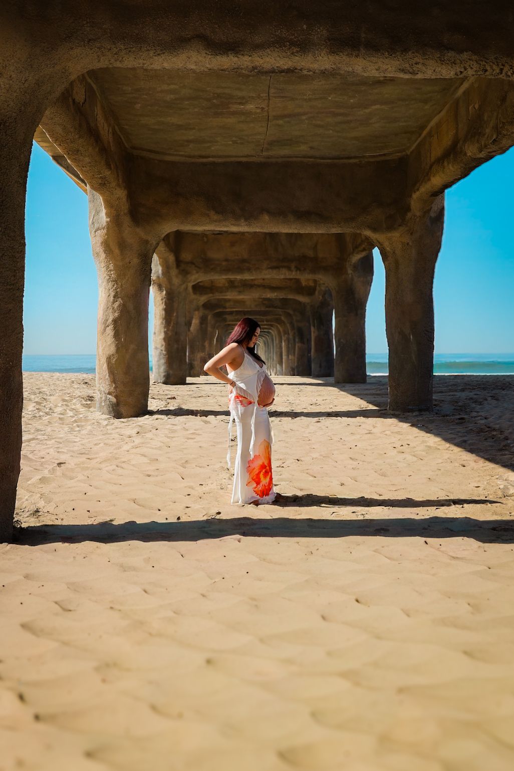 Side profile of pregnant woman in a white floral two-piece on the beach, sunlight highlighting her belly and flowing skirt.