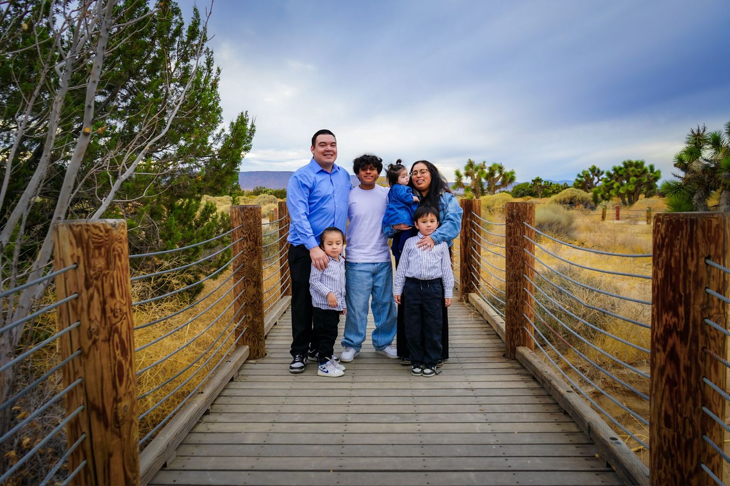 Family of six standing on a wood bridge surrounded with a desert terrain and joshua trees, smiling and holding onto each other.