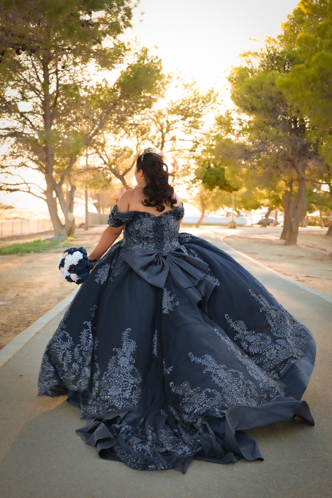 Girl in a blue quinceañera dress walking down a path towards the sunset with the warm glow illuminating her dress.