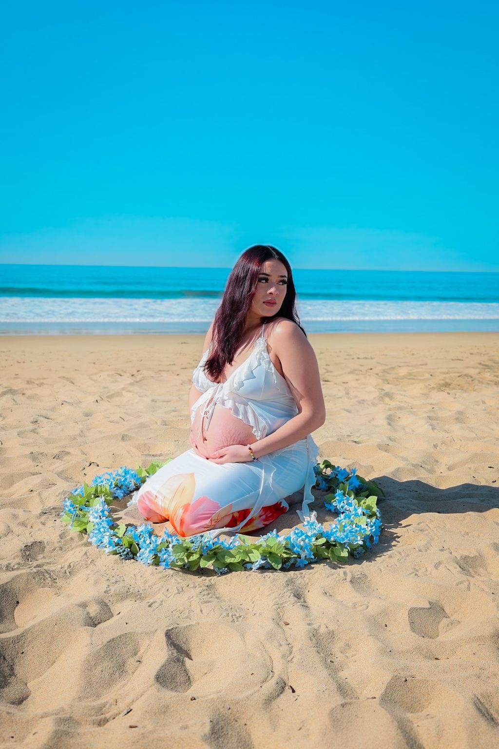 Kneeling maternity portrait of expecting mom in white outfit, surrounded by a delicate blue floral garland on a sunny beach.