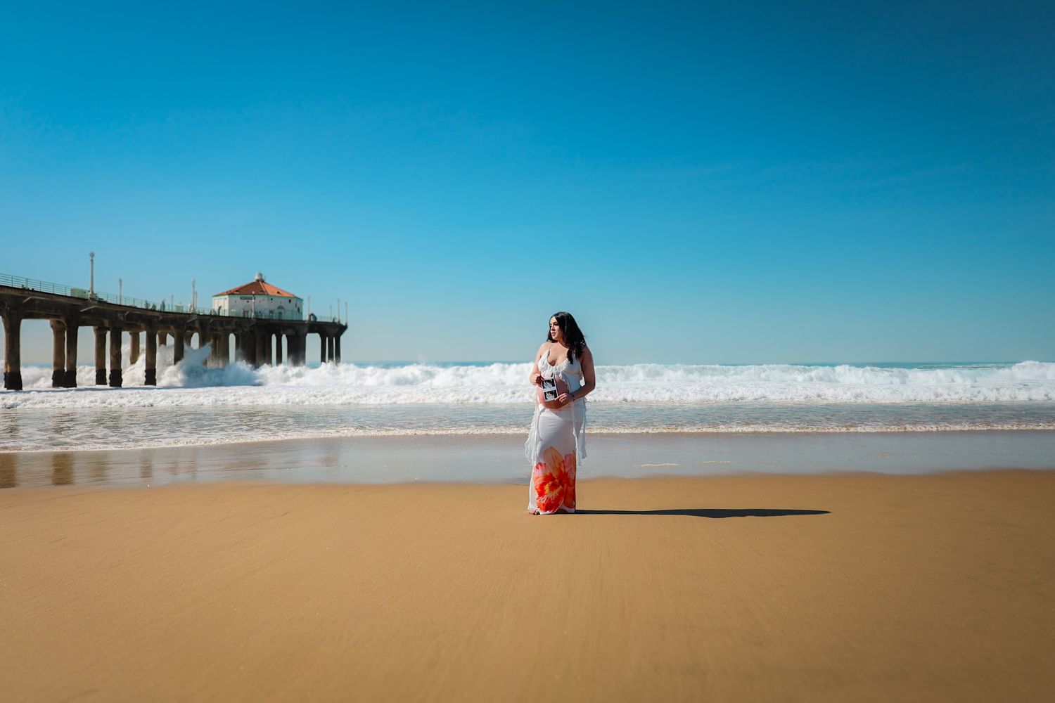 Pregnant mom in a white two-piece outfit with orange floral patterns standing on a sunlit beach during a daytime maternity session.