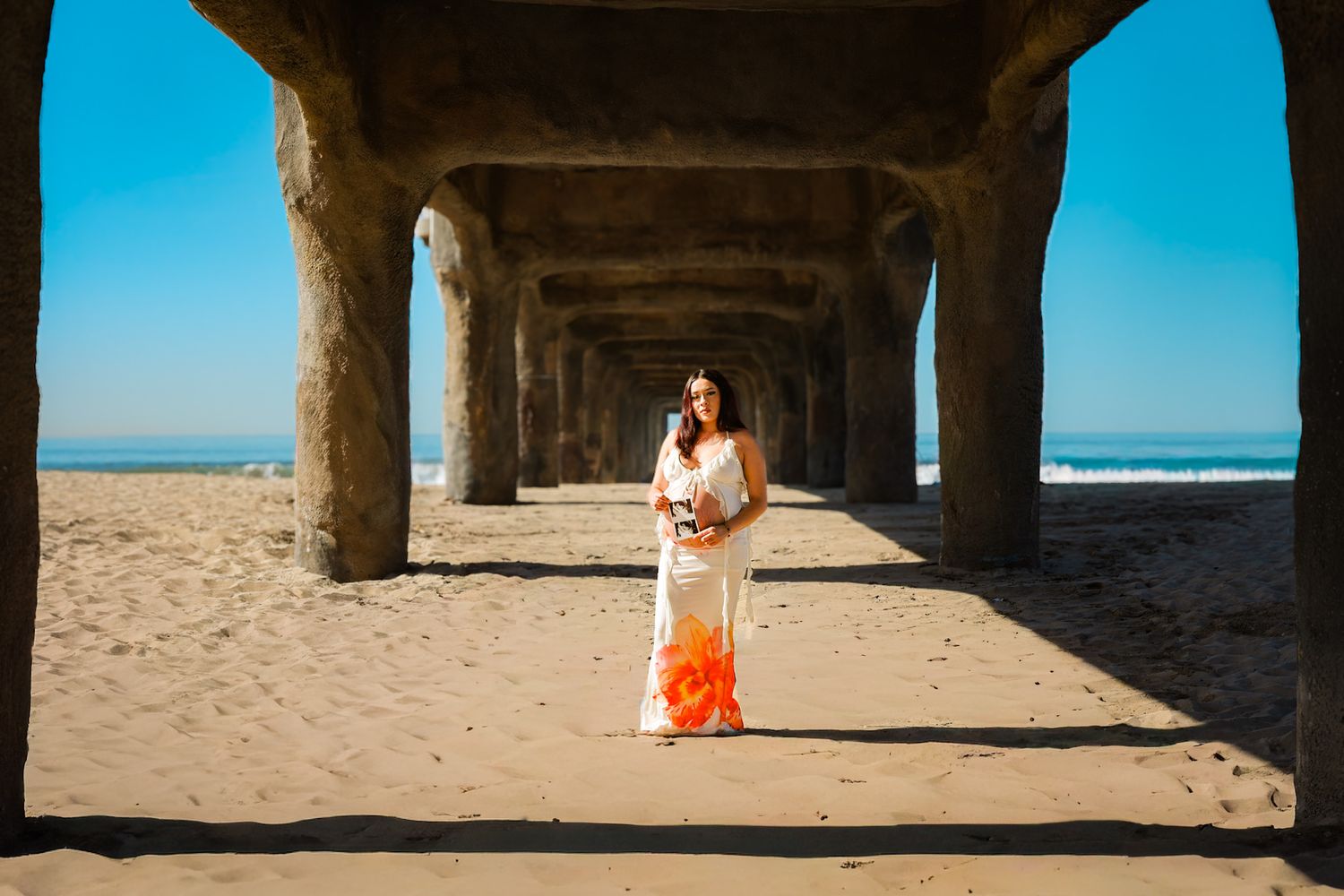 Pregnant woman in a white and orange floral two-piece posing on a sunny beach, celebrating her maternity glow.