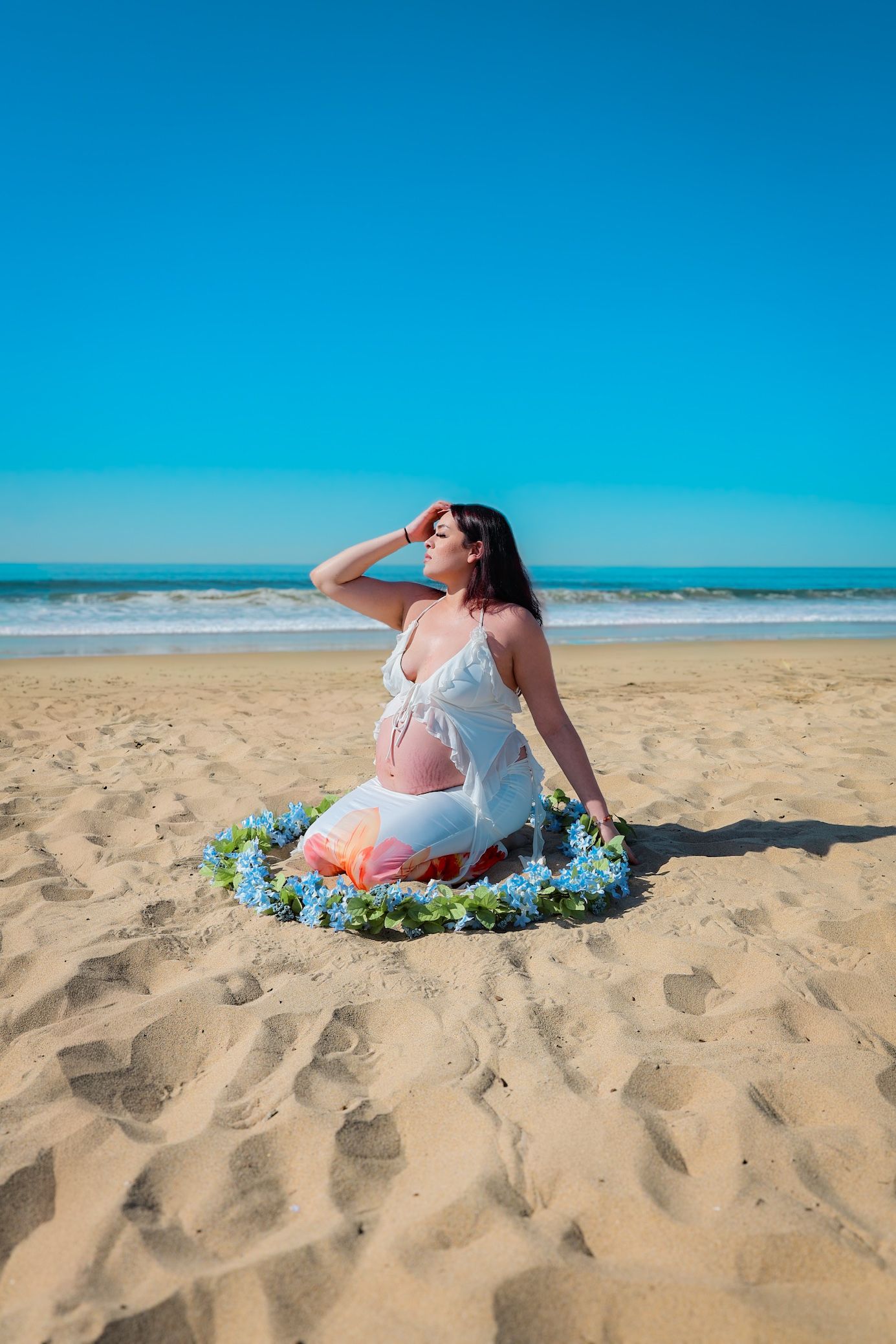 Expecting mother kneeling on sunlit sand in white two-piece, encircled by a blue floral garland during a maternity session.