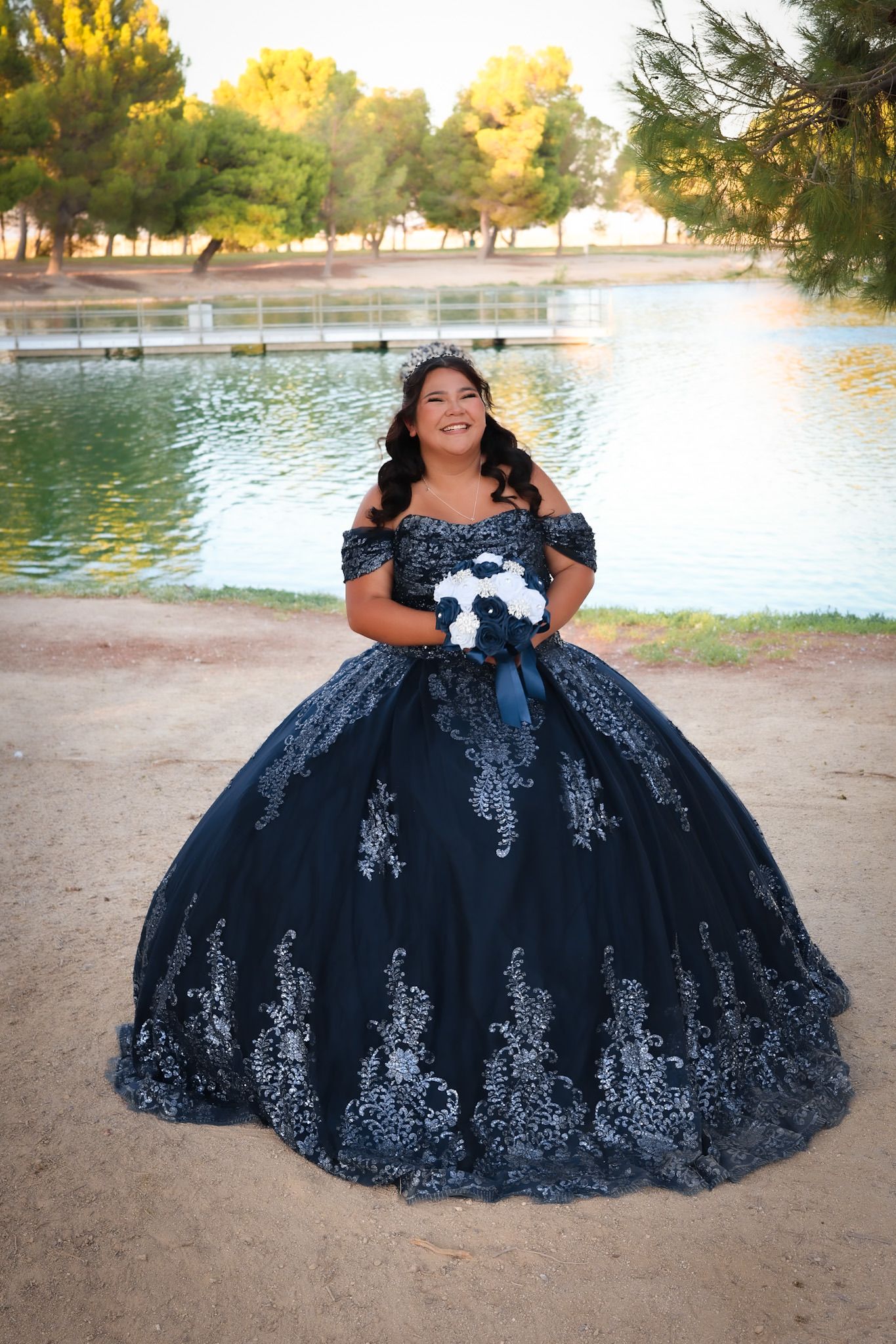 Girl in a blue quinceañera dress posing near a lake before her celebration, sunlight reflecting off the water and highlighting her gown.