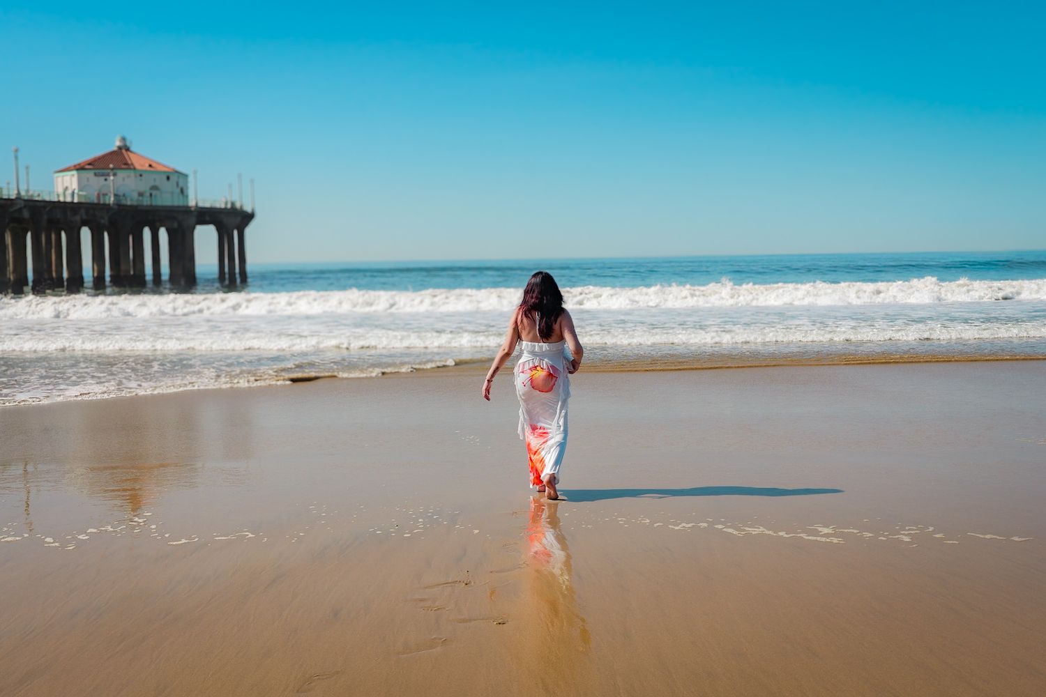 Pregnant mom in a flowing white and orange floral two-piece walking towards the water, looking off into the distance during daytime session.