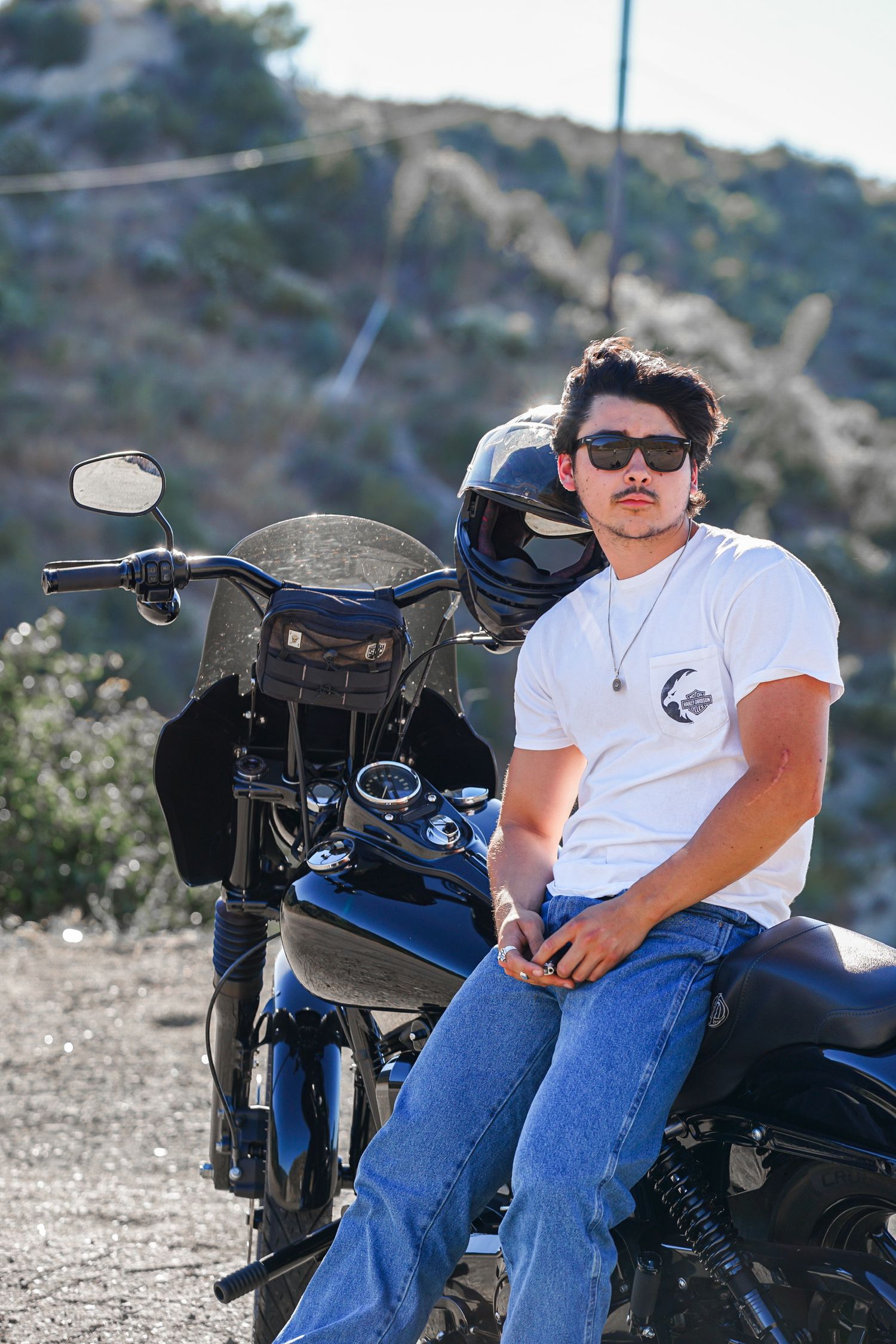 Solo motorcycle rider leaning against his black Harley Davidson, the wind subtly blowing his hair away from his face as he looks away.