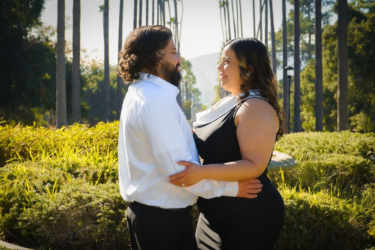 Standing amongst lush greenery, the engaged couple faces each other, smiling softly holding each others waist.