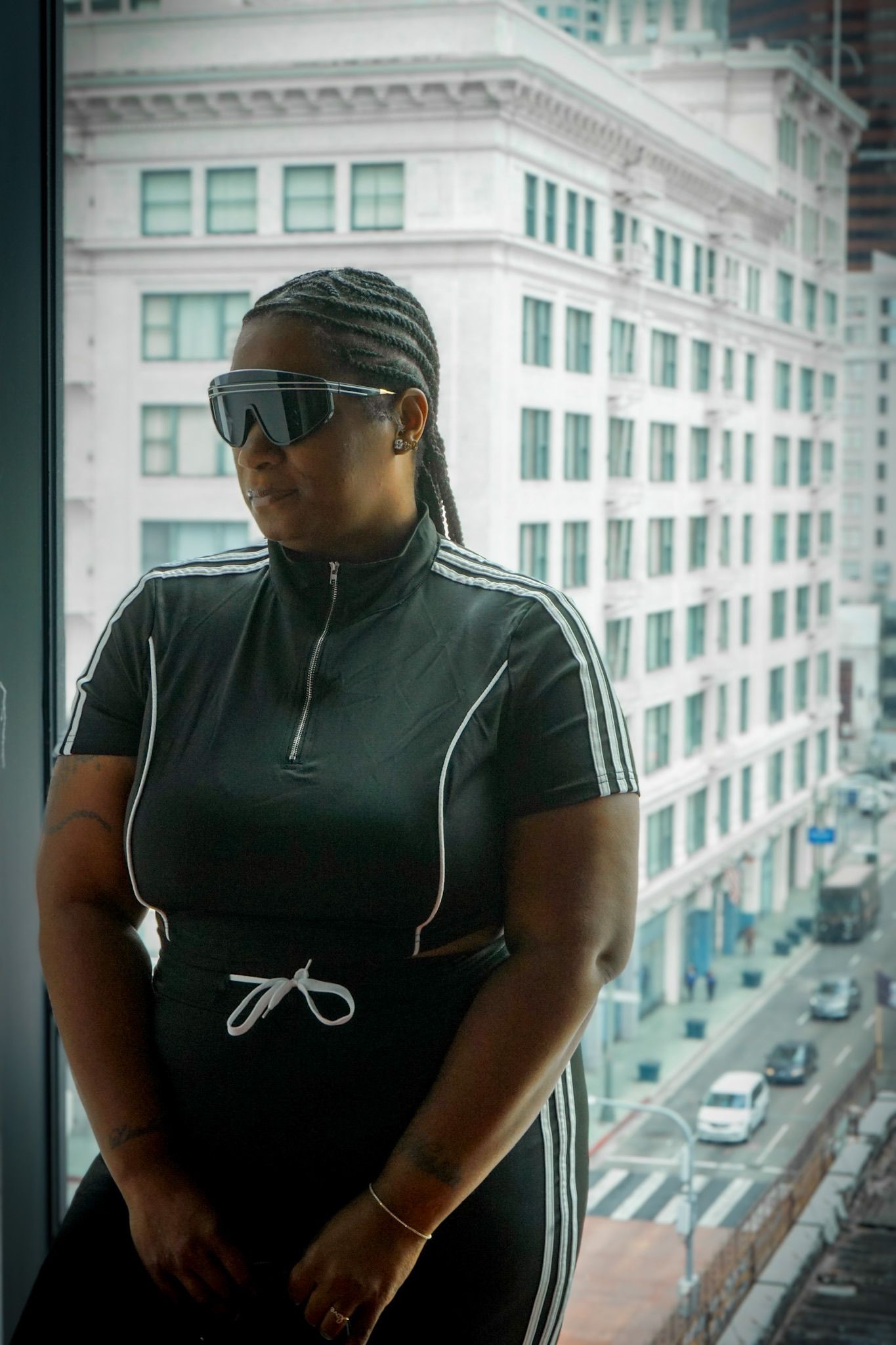 Woman leaning her back against a large glass window with view of the city below, posed, with natural light illuminating her frame.