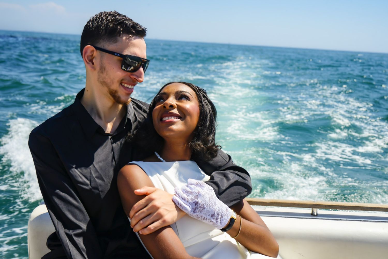 Engaged couple laughing together staring at one another lovingly during engagement shoot while riding in the back of a speed boat.