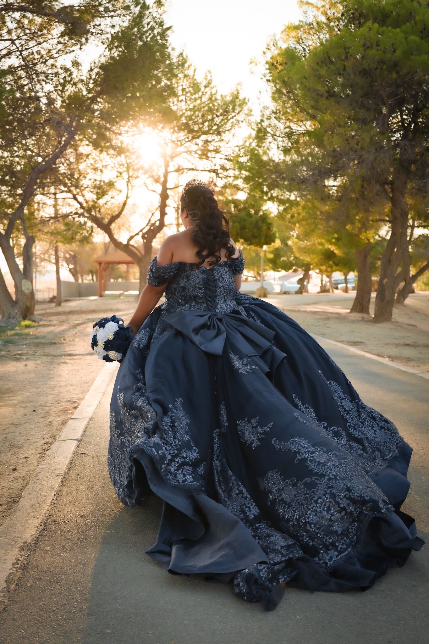 Girl in a blue quinceañera dress walking down a path towards the sunset with the warm glow illuminating her dress.
