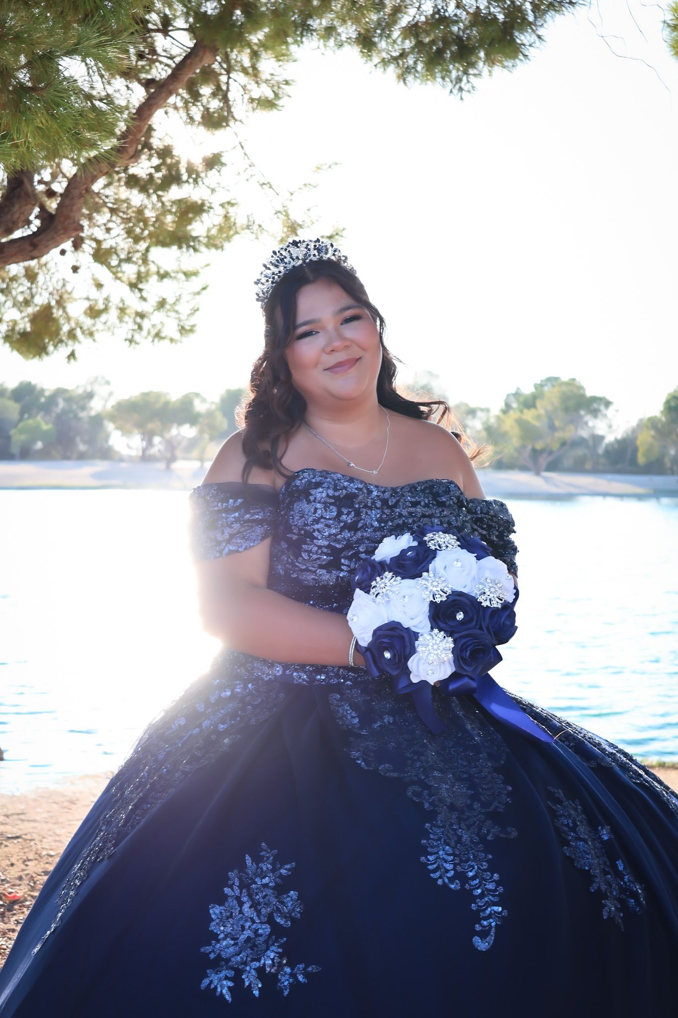Girl in a blue quinceañera dress posing near a lake before her celebration, sunlight reflecting off the water and highlighting her gown.