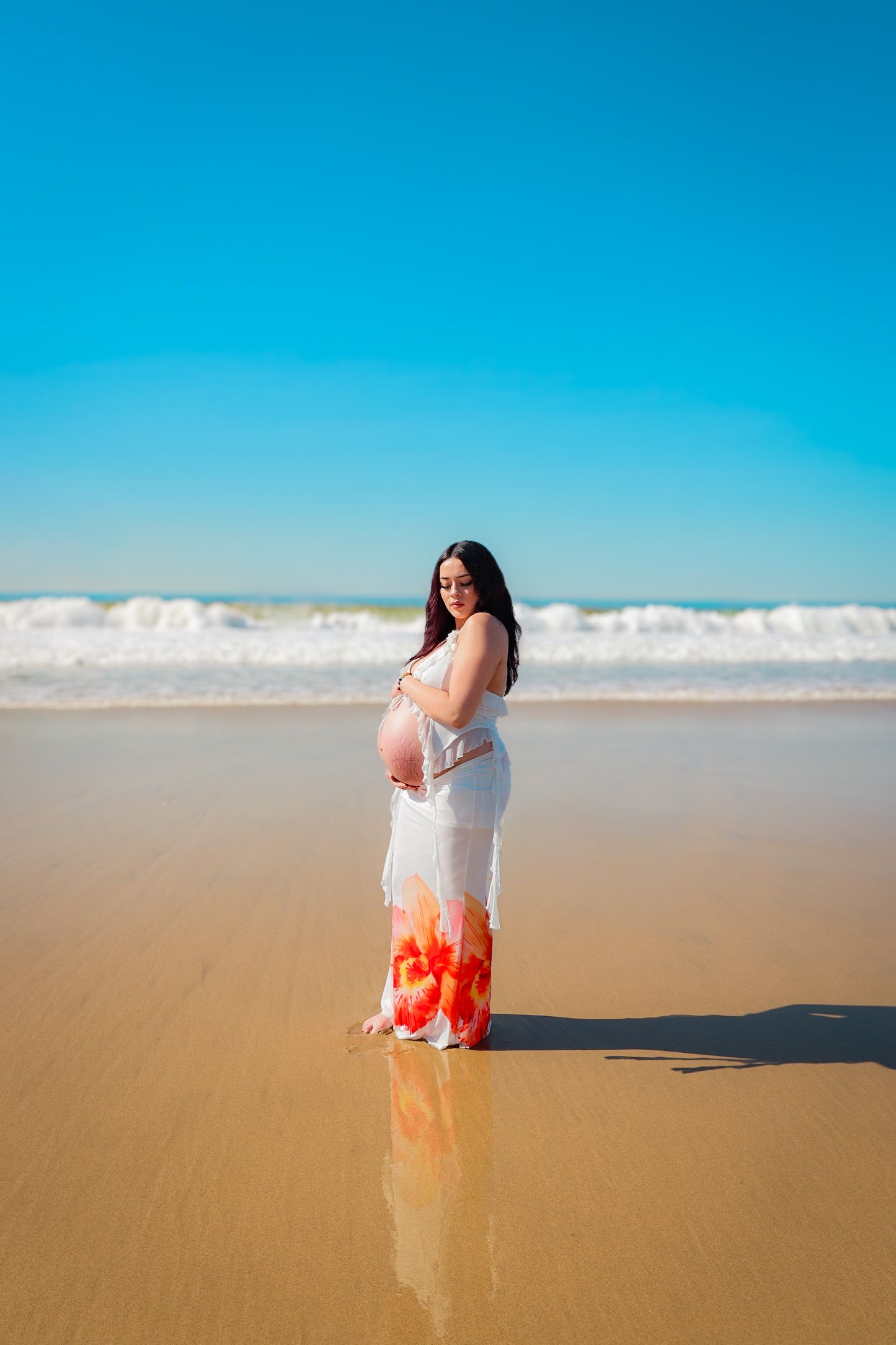 Pregnant woman in a white and orange floral two-piece posing on a sunny beach, celebrating her maternity glow caressing her belly.