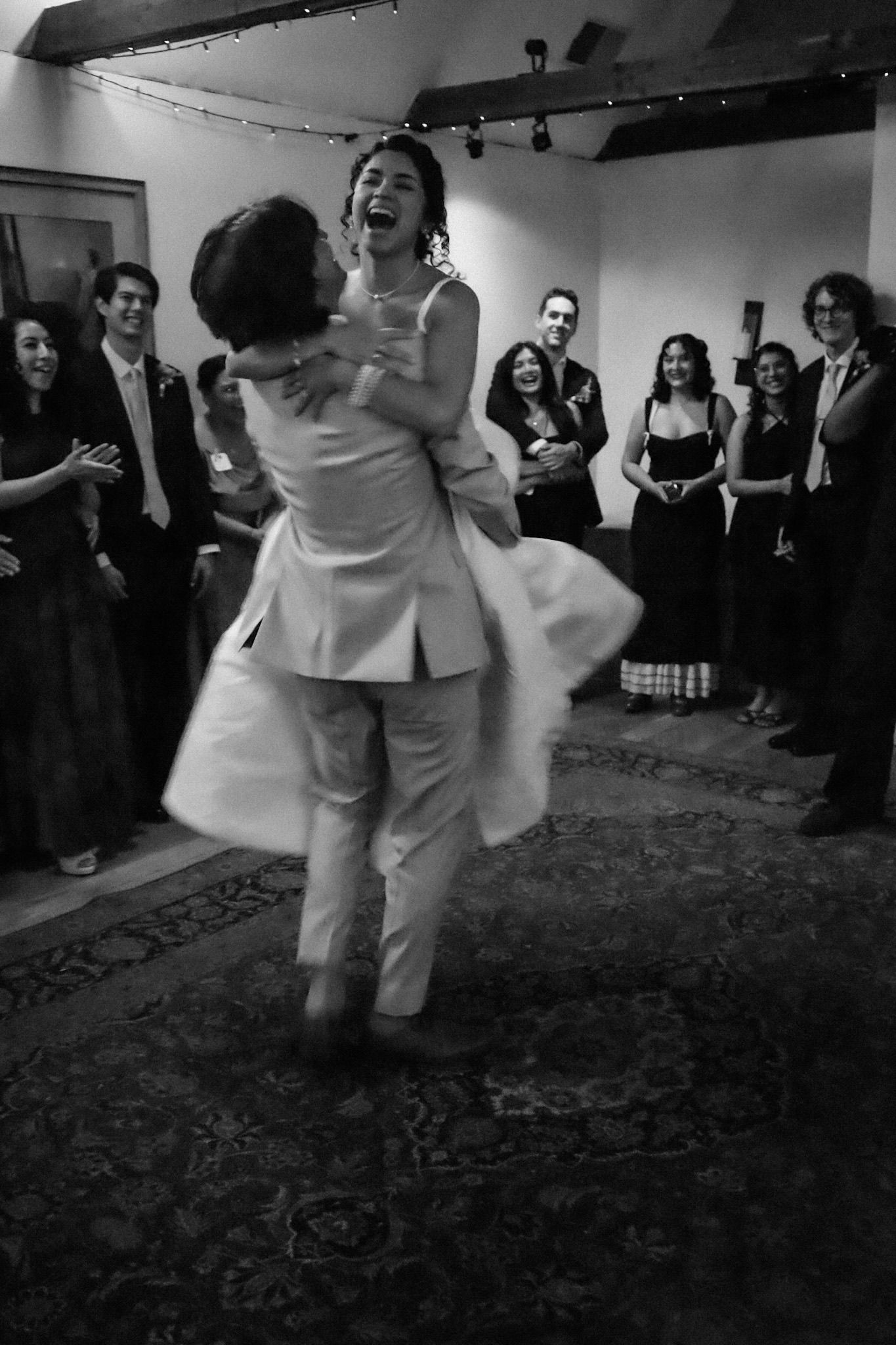 Black and white still of newlywed couple with the groom twirling the bride around in a circle with family and friends onlooking in joy.