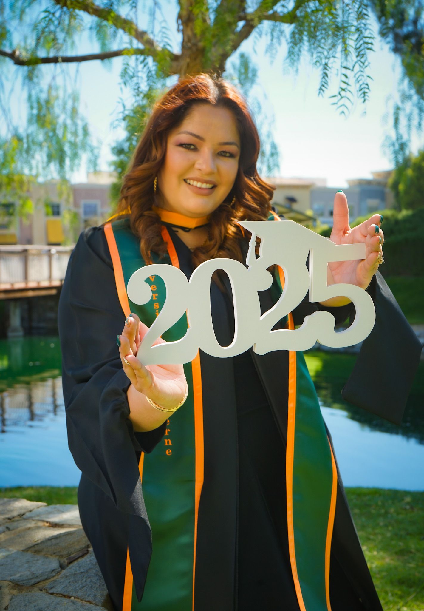 2025 college graduate golding a white wooden 2025 sign with a graduation cap on the number 2, smiling and wearing her robe and sash.