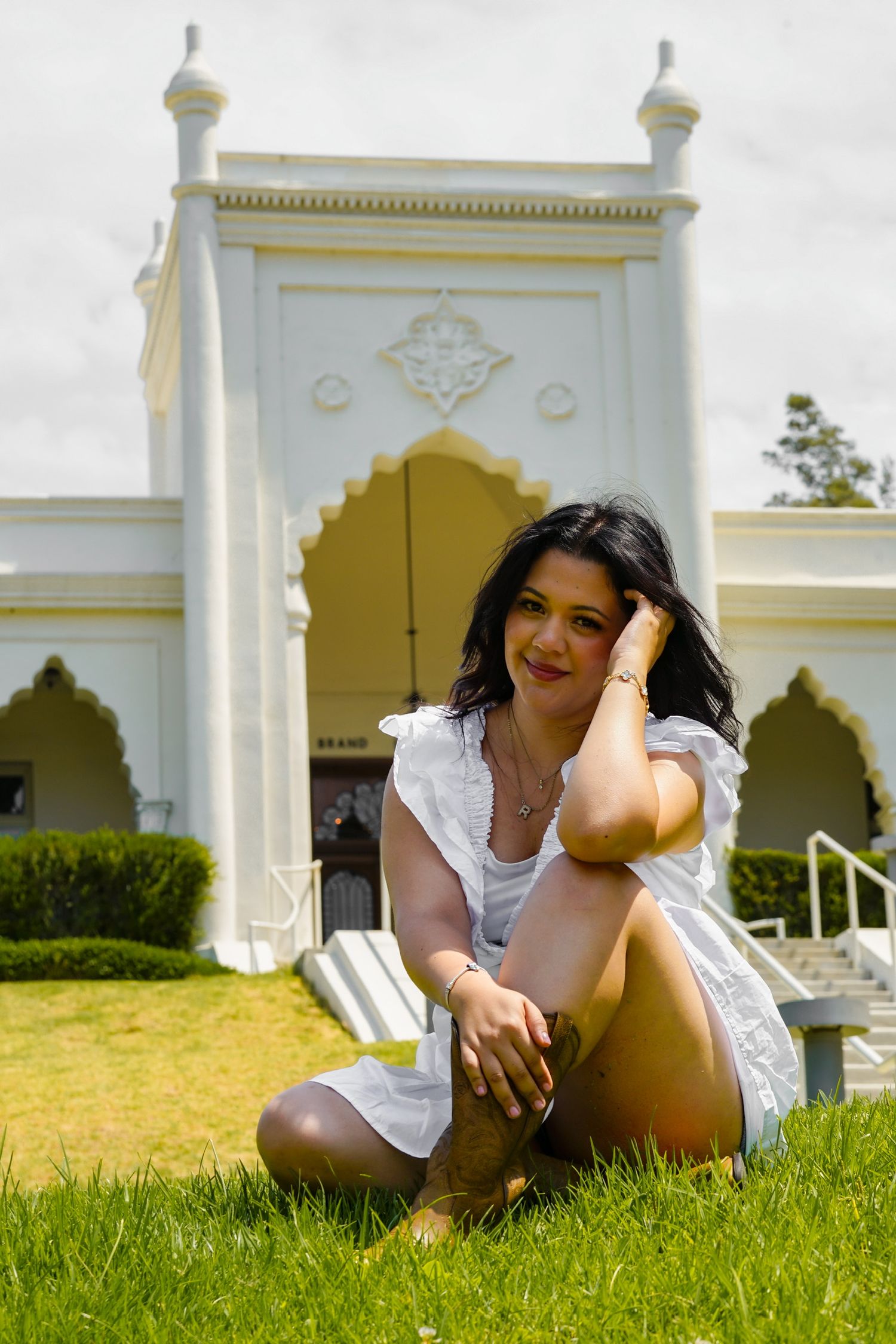 Woman sitting in front of a white library with beautiful architecture, wearing a flowing white dress and cowboy boots smiling softly.