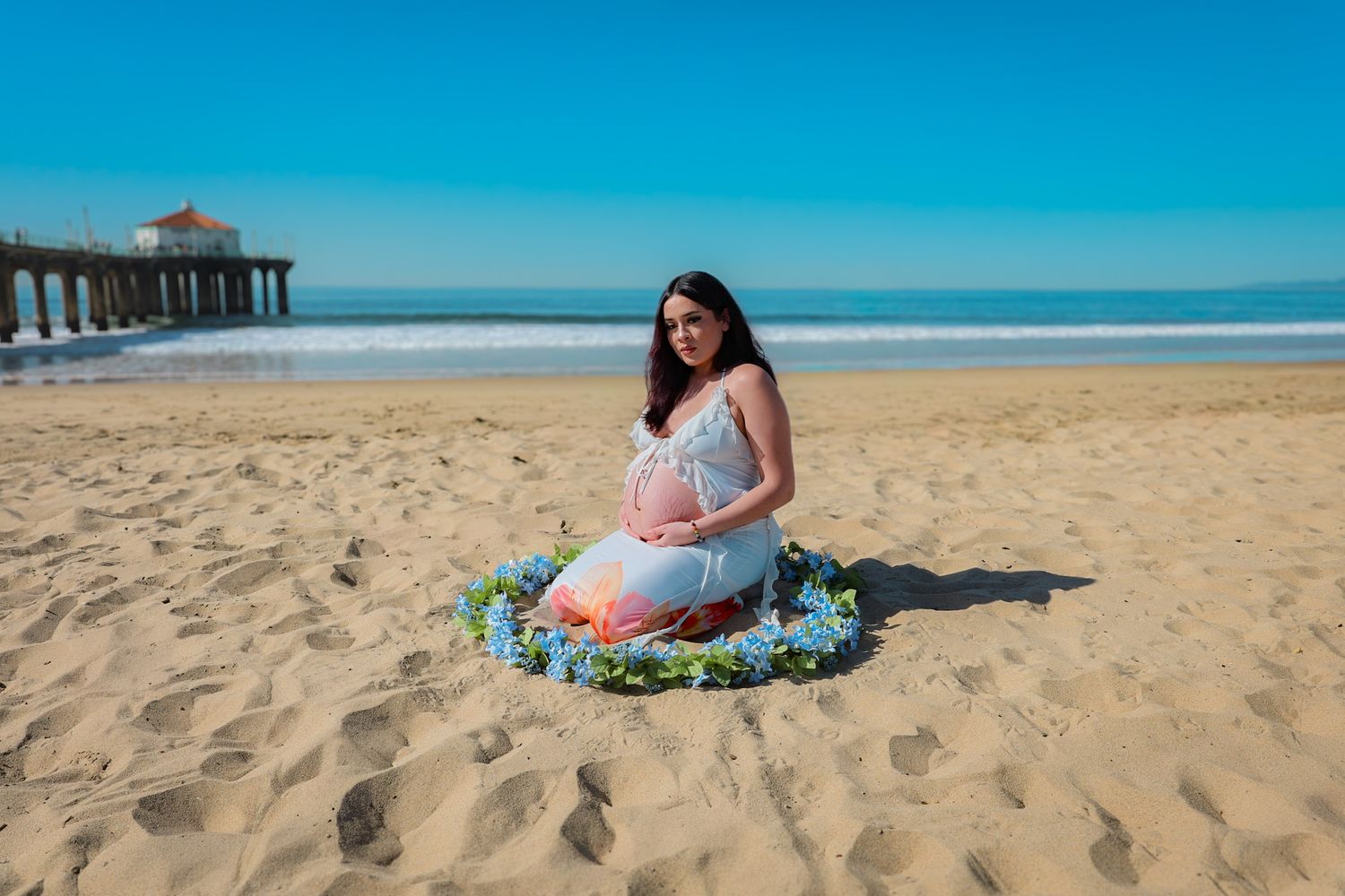 Kneeling maternity portrait of expecting mom in white outfit, surrounded by a delicate blue floral garland on a sunny beach.