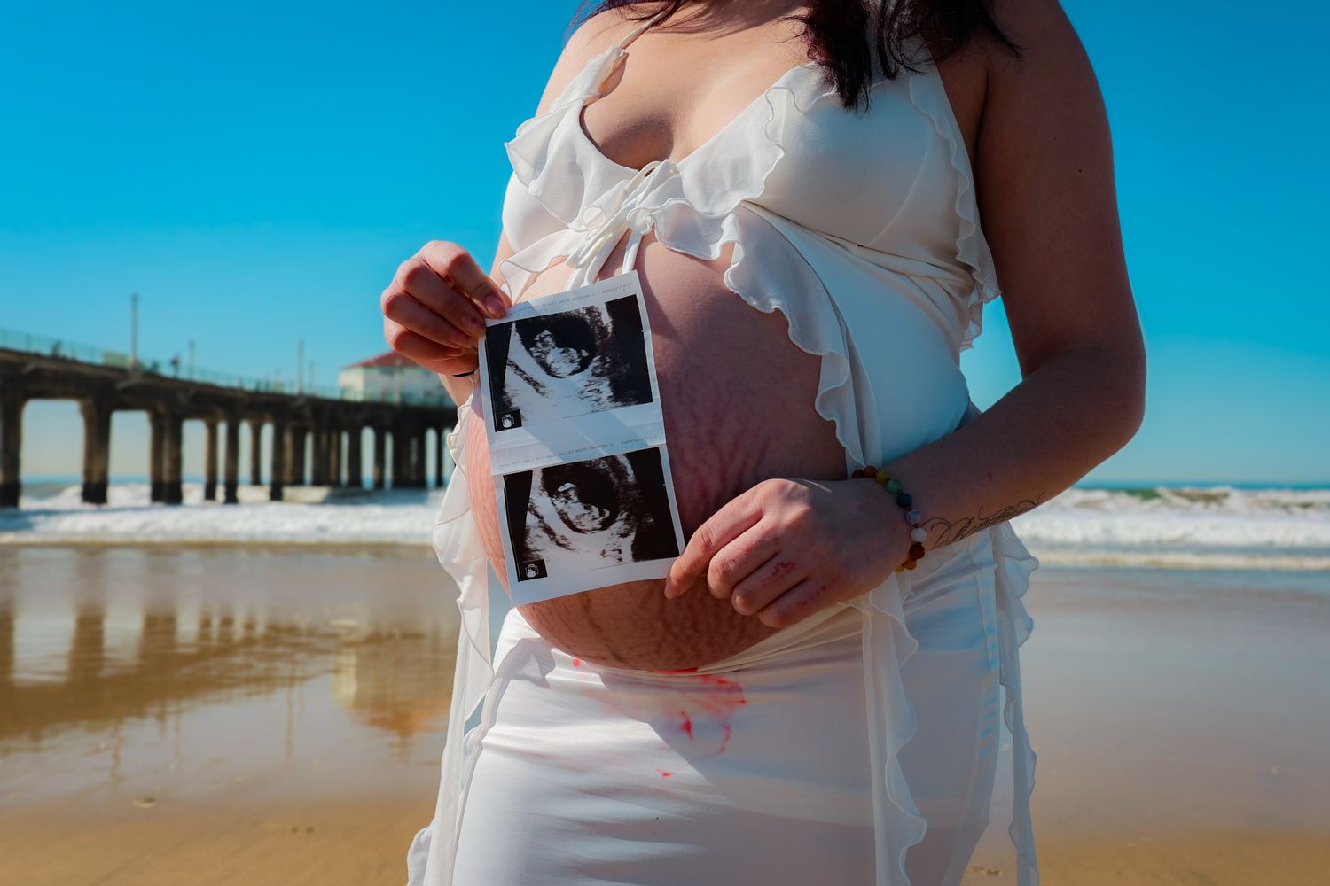 Close-up of pregnant woman in white floral two-piece on sunlit beach, holding her ultrasound image.