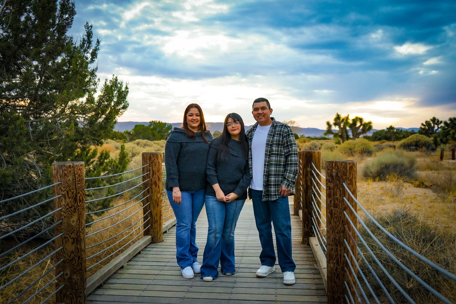 Family of three smiling warmly surrounded by desert foliage, a beautiful cloudy sky with hints of sunlight breaking through.